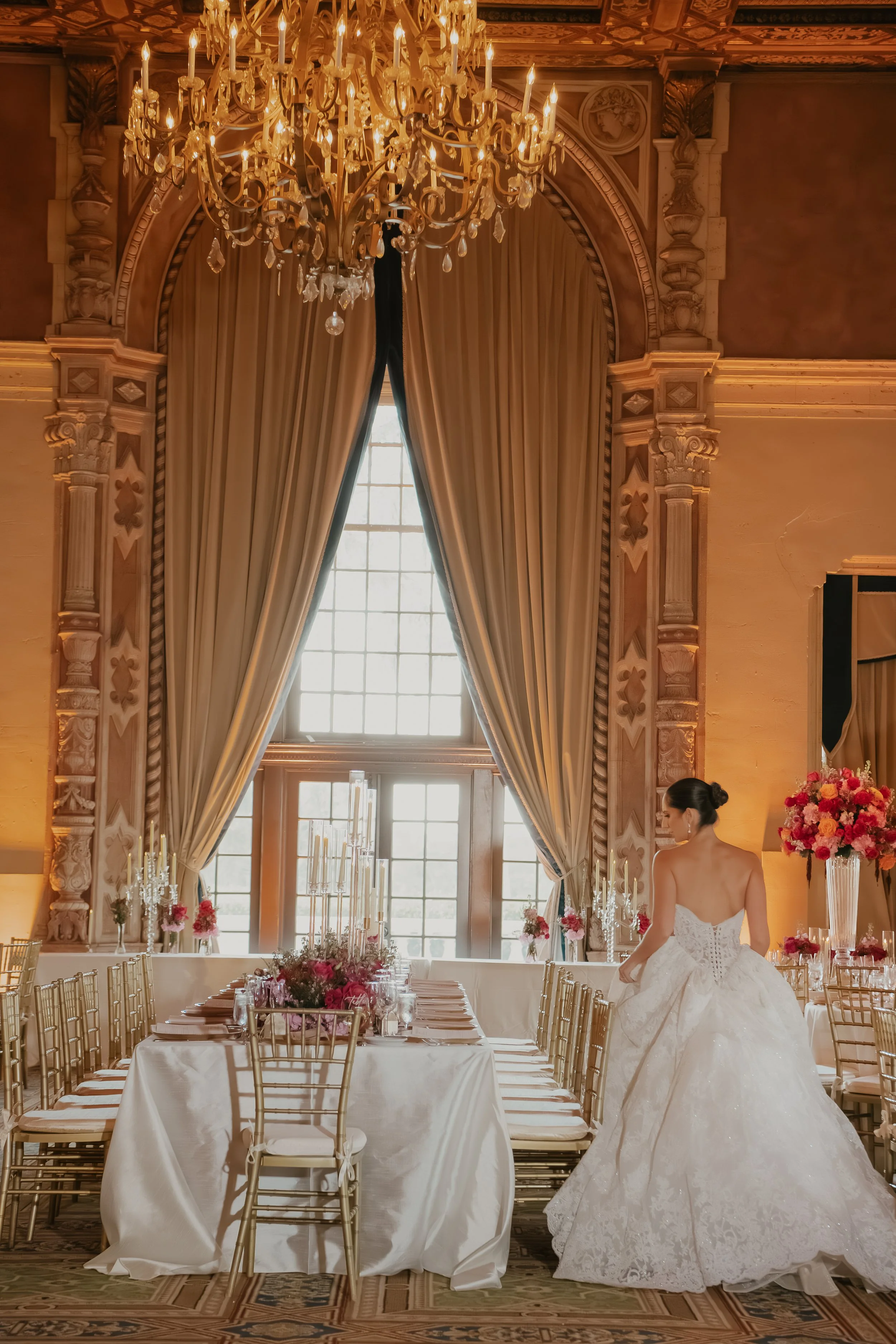 Elegant wedding reception room with long table set with floral centerpieces and gold chairs, large window with draped curtains, ornate chandelier, and a bride in a white wedding gown.