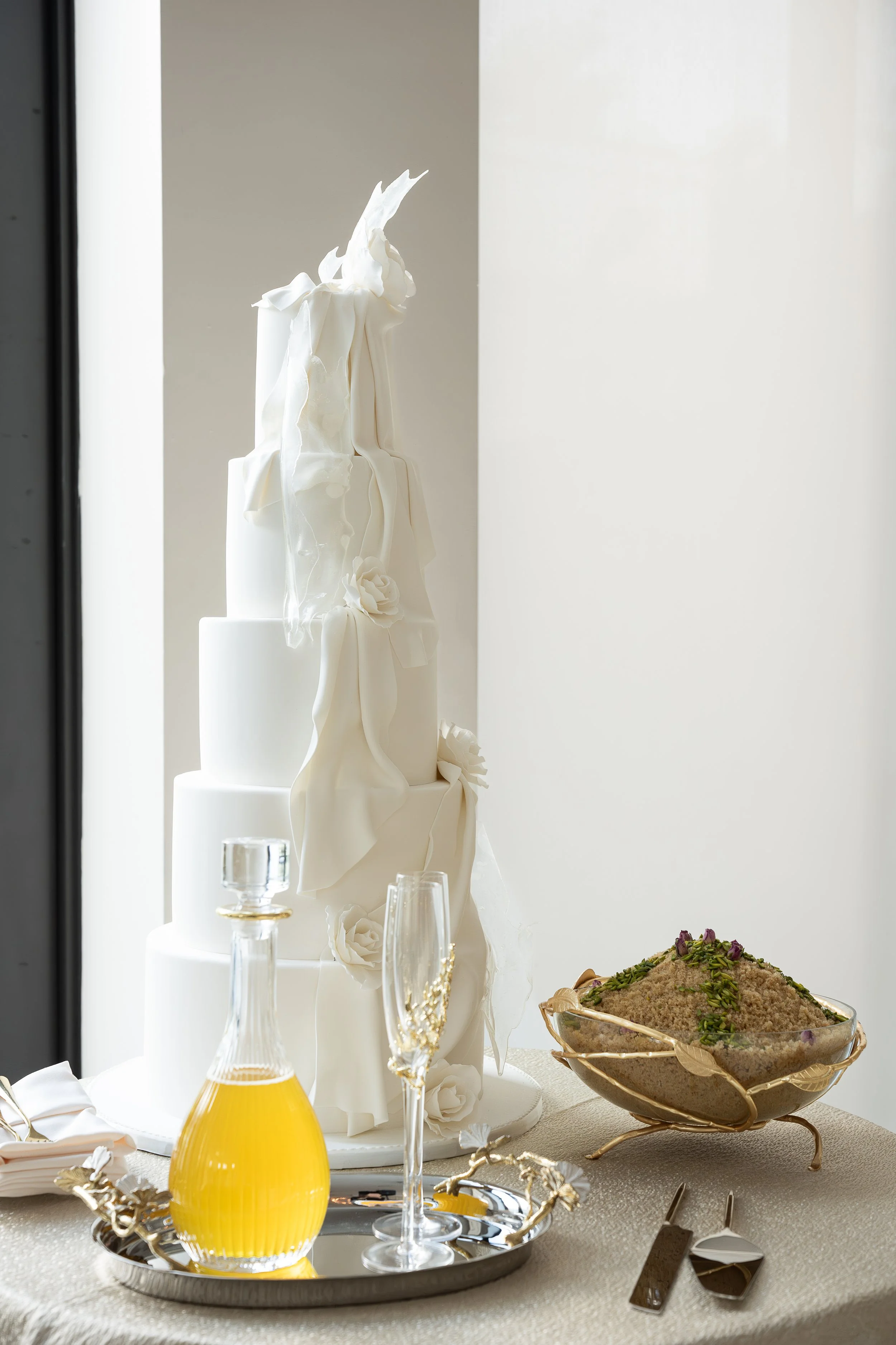 A table set with a white wedding cake, champagne glasses, a yellow drink, and a bowl of dessert garnished with pistachios and rose petals.