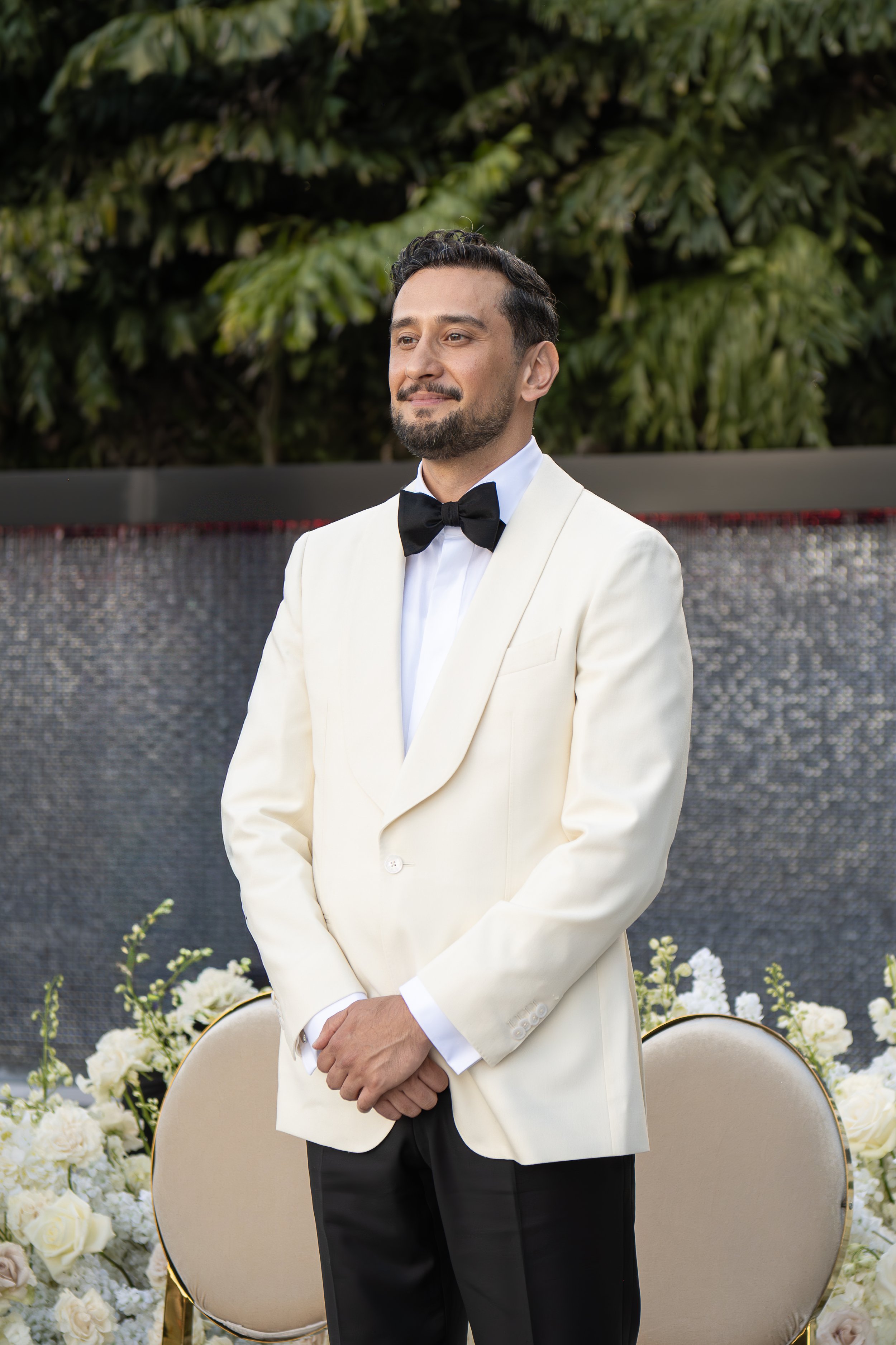 A man in a white tuxedo with a black bowtie standing at a formal event, with greenery and white flowers in the background.