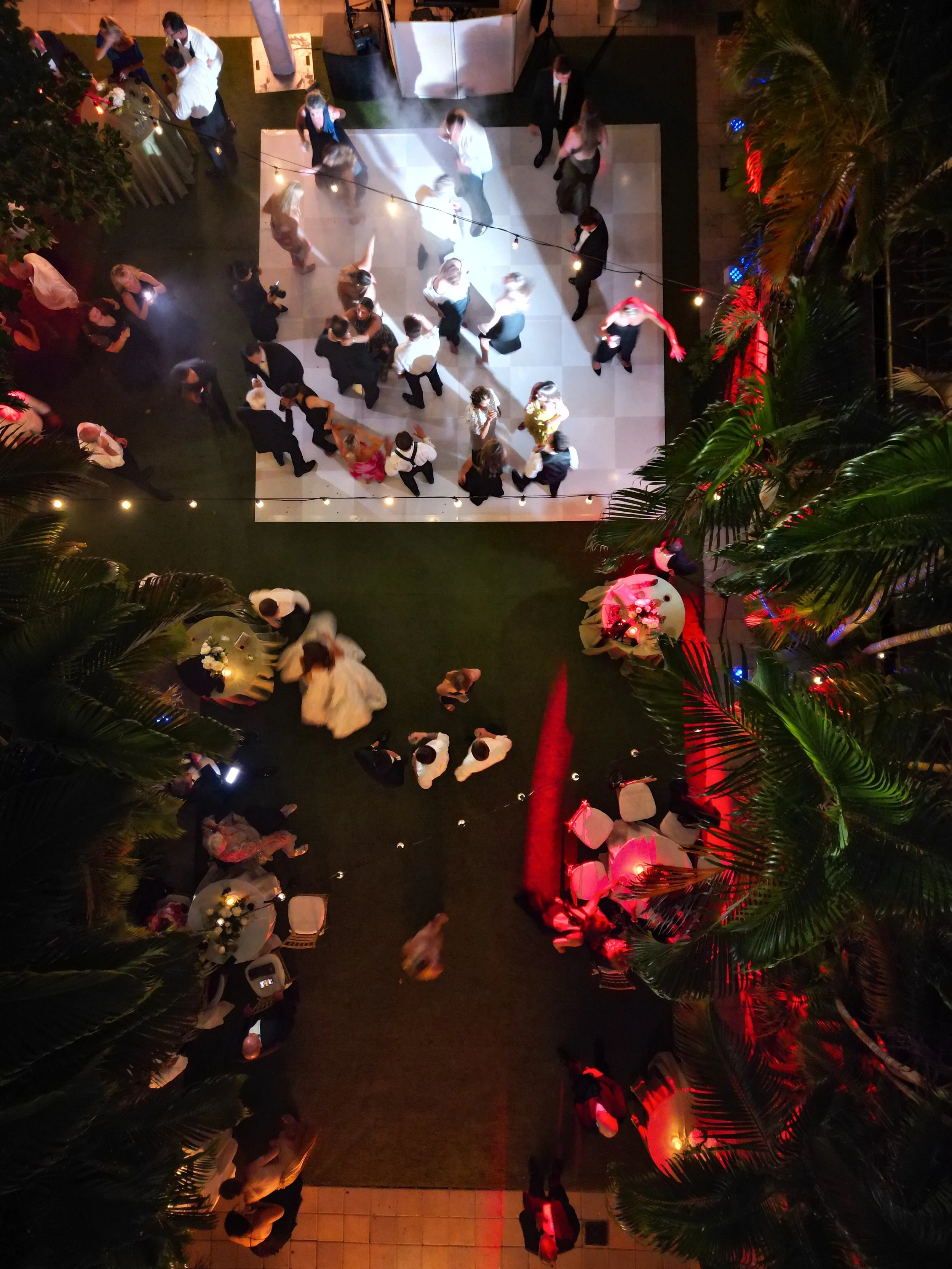 People dancing on a checkered dance floor at a social event with string lights. Lush green palm trees frame the scene, which includes tables with floral centerpieces, decorations, and a DJ booth.