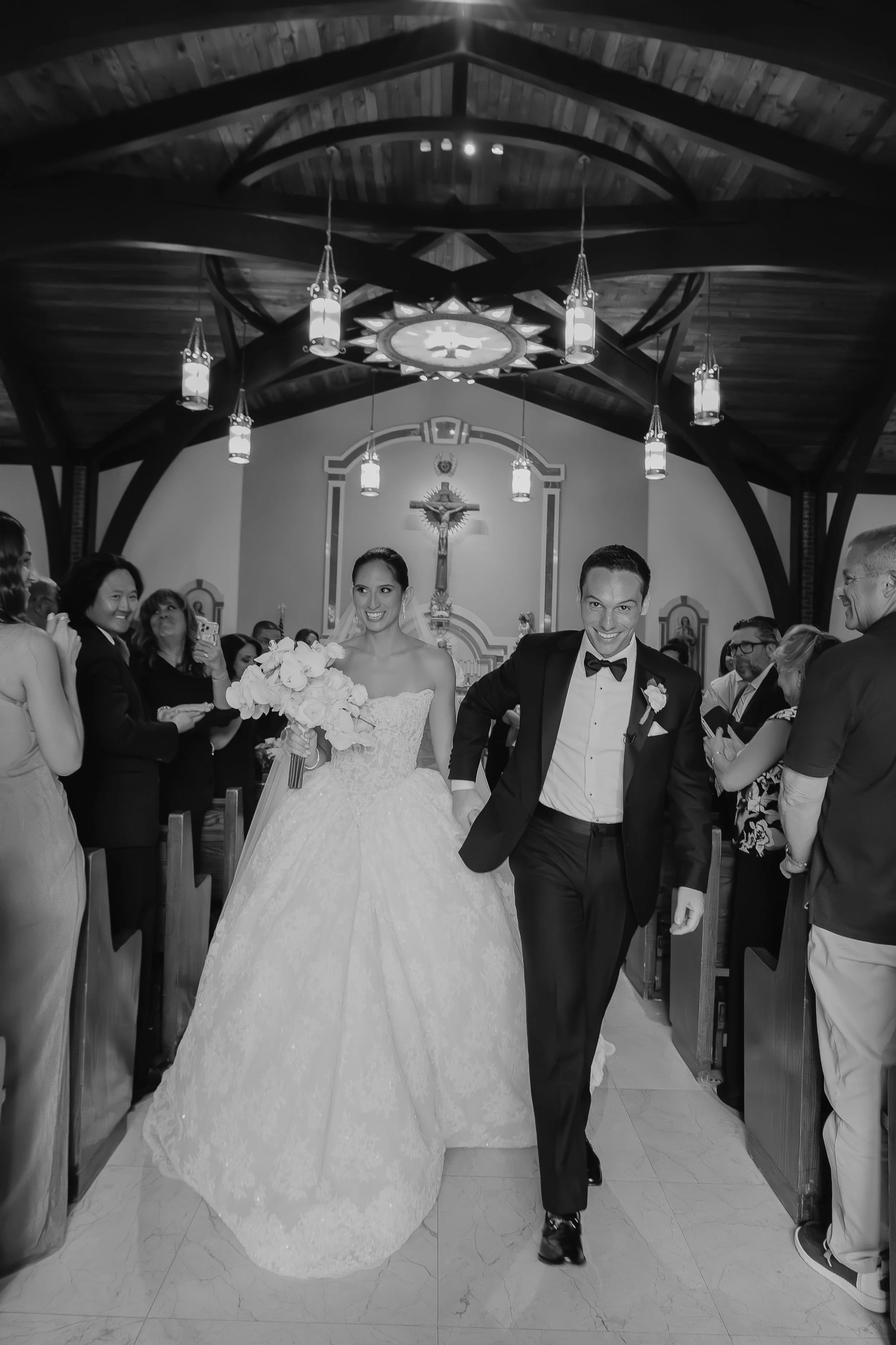 A bride and groom walking down the aisle after their wedding ceremony inside a church, surrounded by guests and wedding attendants, with a crucifix on the altar in the background.
