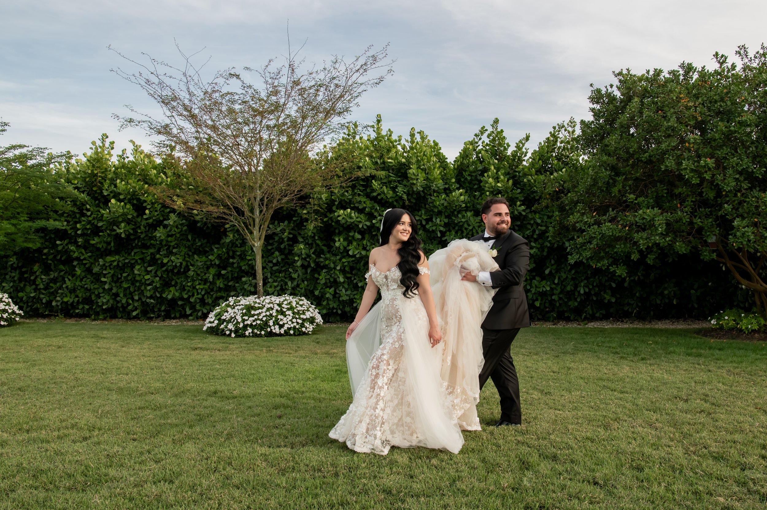 A newlywed couple walking on a grassy lawn in front of lush green bushes, with the groom holding the bride's train, both smiling.