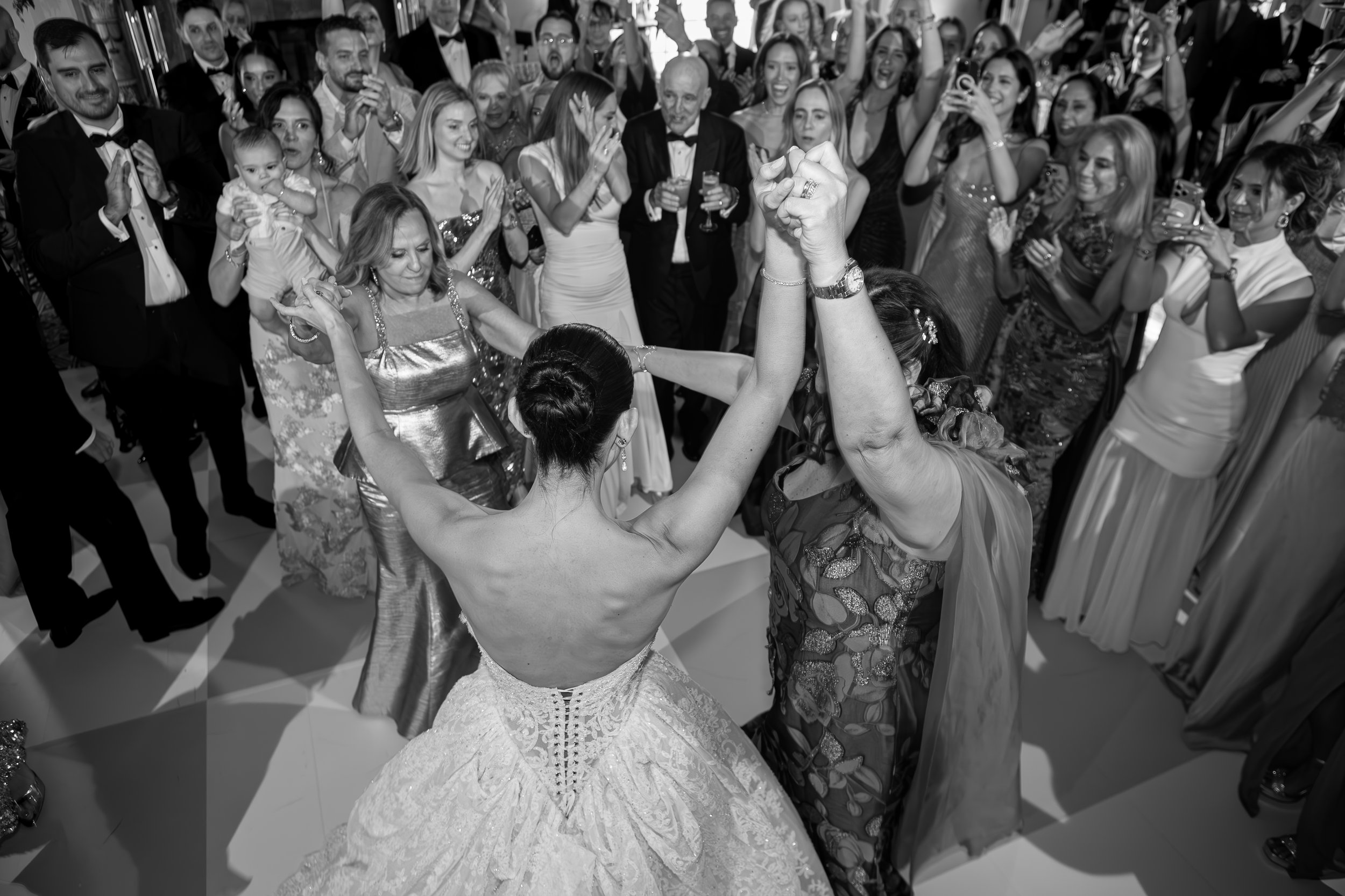 A large group of people dressed in formal attire celebrating, with some dancing and others holding drinks, at an indoor event.