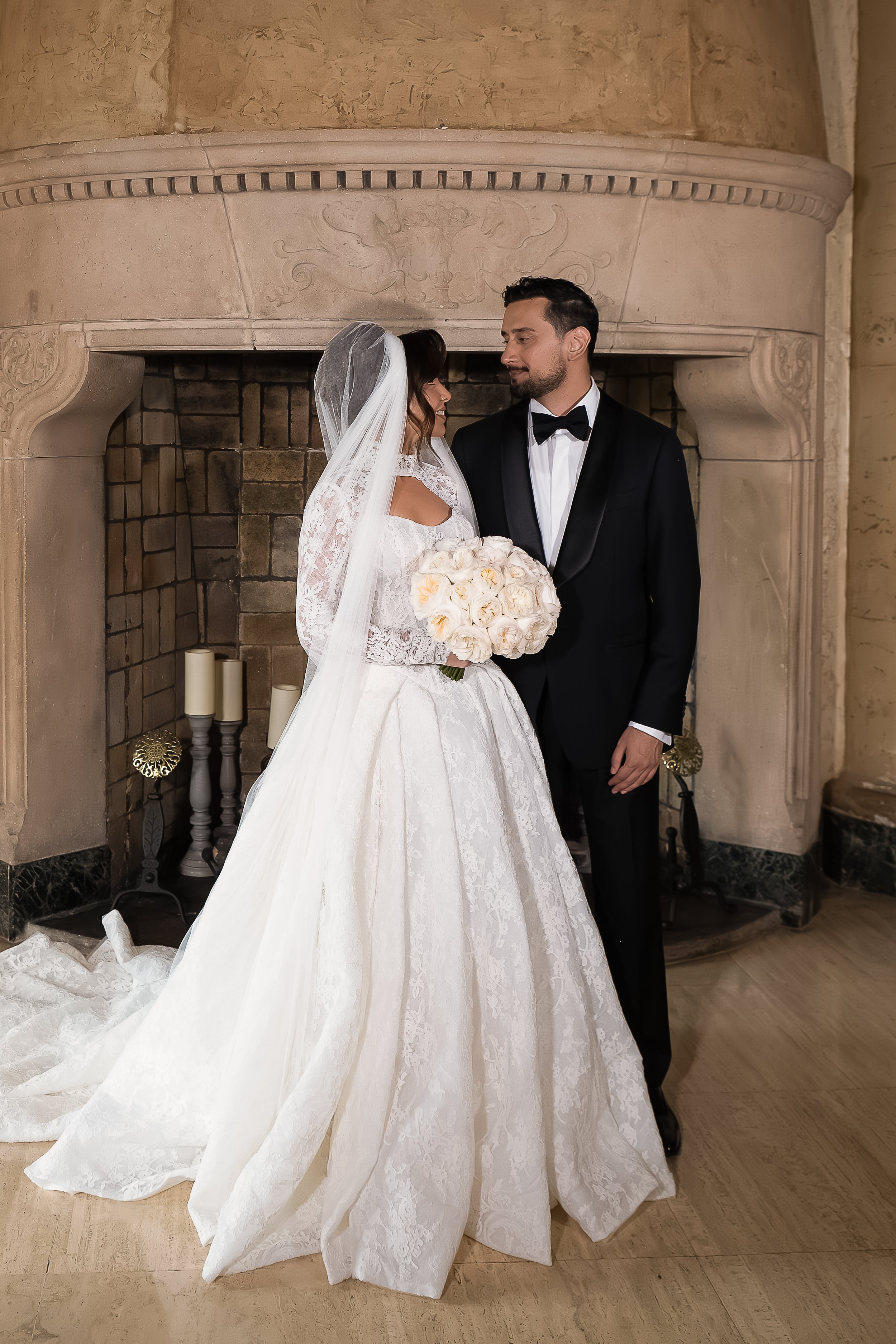 A bride and groom standing in front of a fireplace, looking at each other. The bride is wearing a white lace wedding dress with a long train and veil, holding a bouquet of white roses. The groom is dressed in a black tuxedo with a bow tie. The backgr