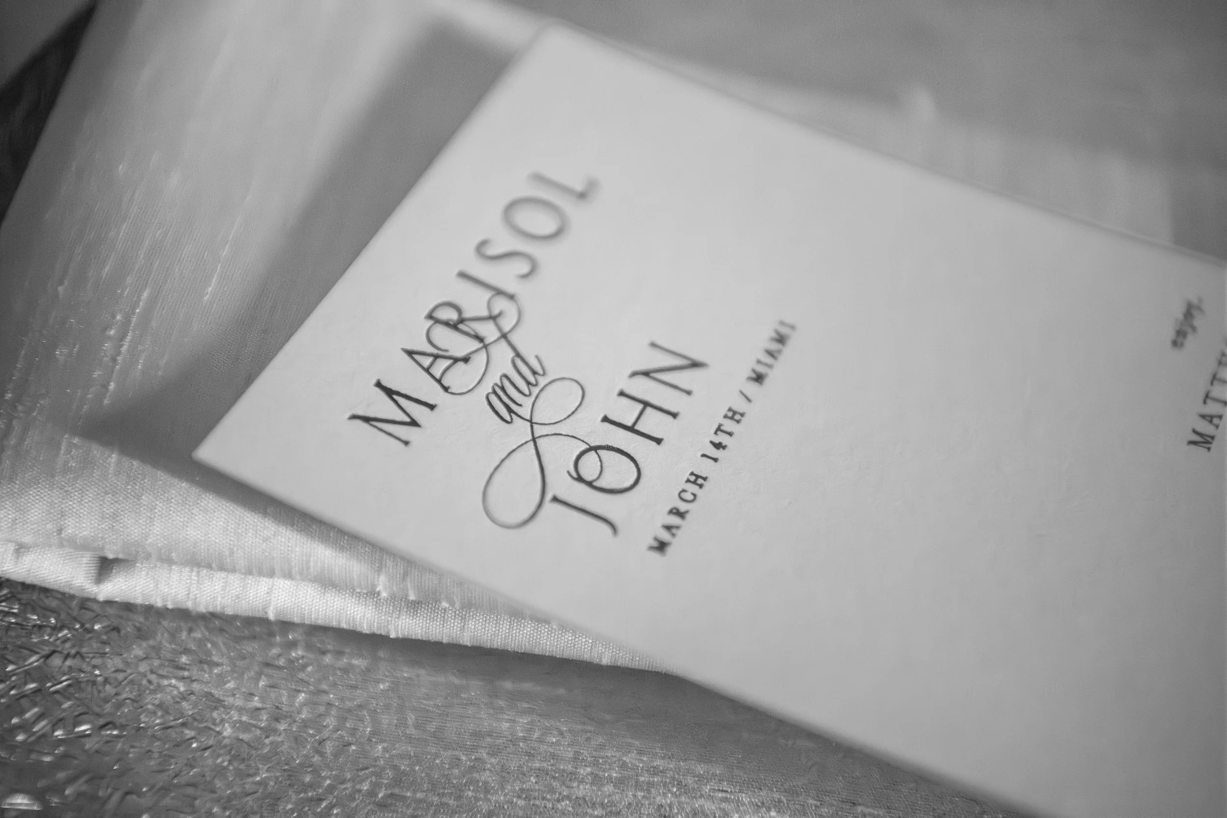 Close-up of a wedding invitation card with elegant scripted and printed text lying on a textured surface. The card features the names 'Marcel & Jane' in decorative script.