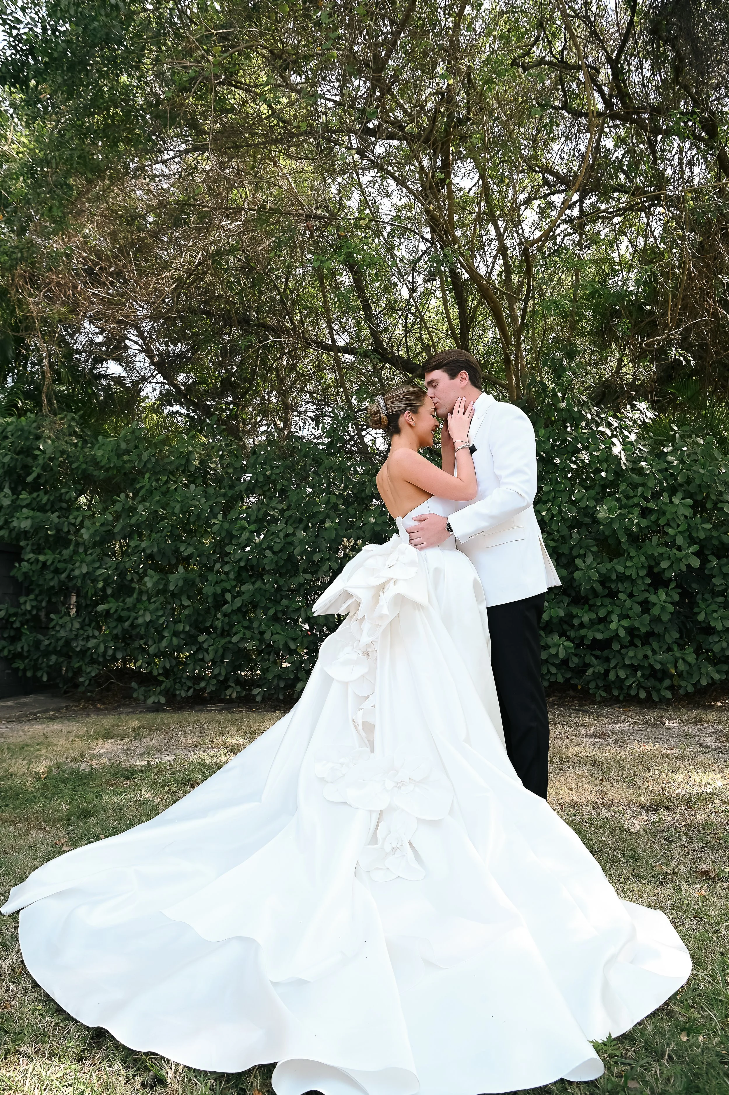 A bride and groom sharing a romantic moment outdoors, with the groom gently touching the bride's face as they nuzzle each other. The bride is wearing a white wedding dress with ruffles and a long train, and the groom is in a white tuxedo jacket with 