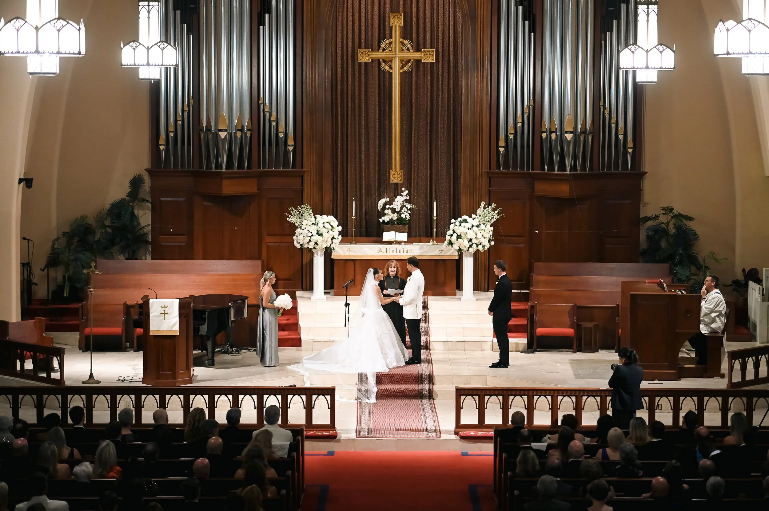 A wedding ceremony taking place inside a church with an altar, a cross, and organ pipes in the background. The bride and groom stand at the altar with an officiant, while a bridesmaid and a groomsman stand nearby. Guests are seated in pews, watching 