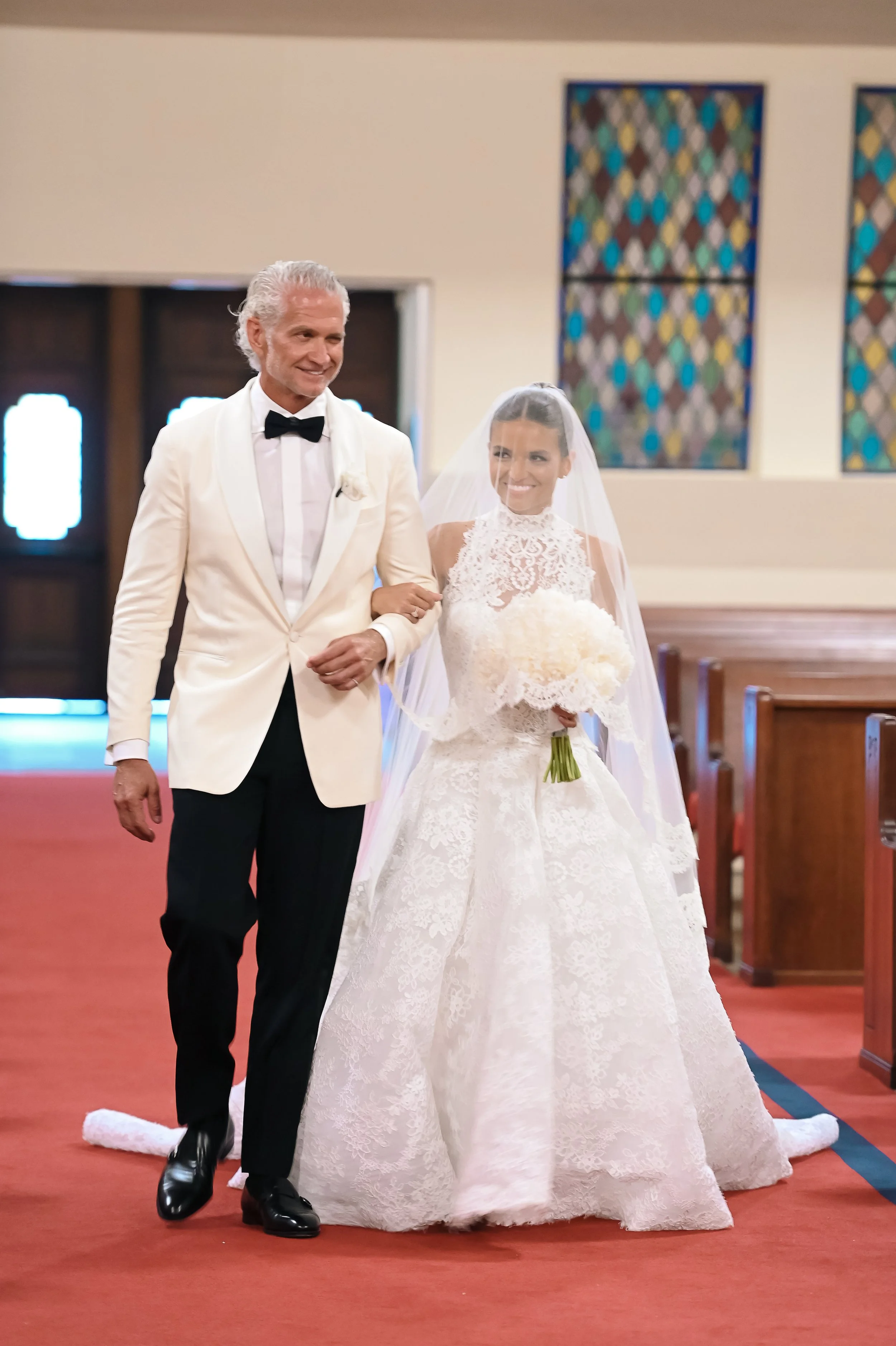A bride and an older man walk together inside a church. The bride wears a white lace wedding gown and veil, holding a bouquet. The man is dressed in a white tuxedo jacket with a black bow tie and black pants. They are smiling and walking down the ais