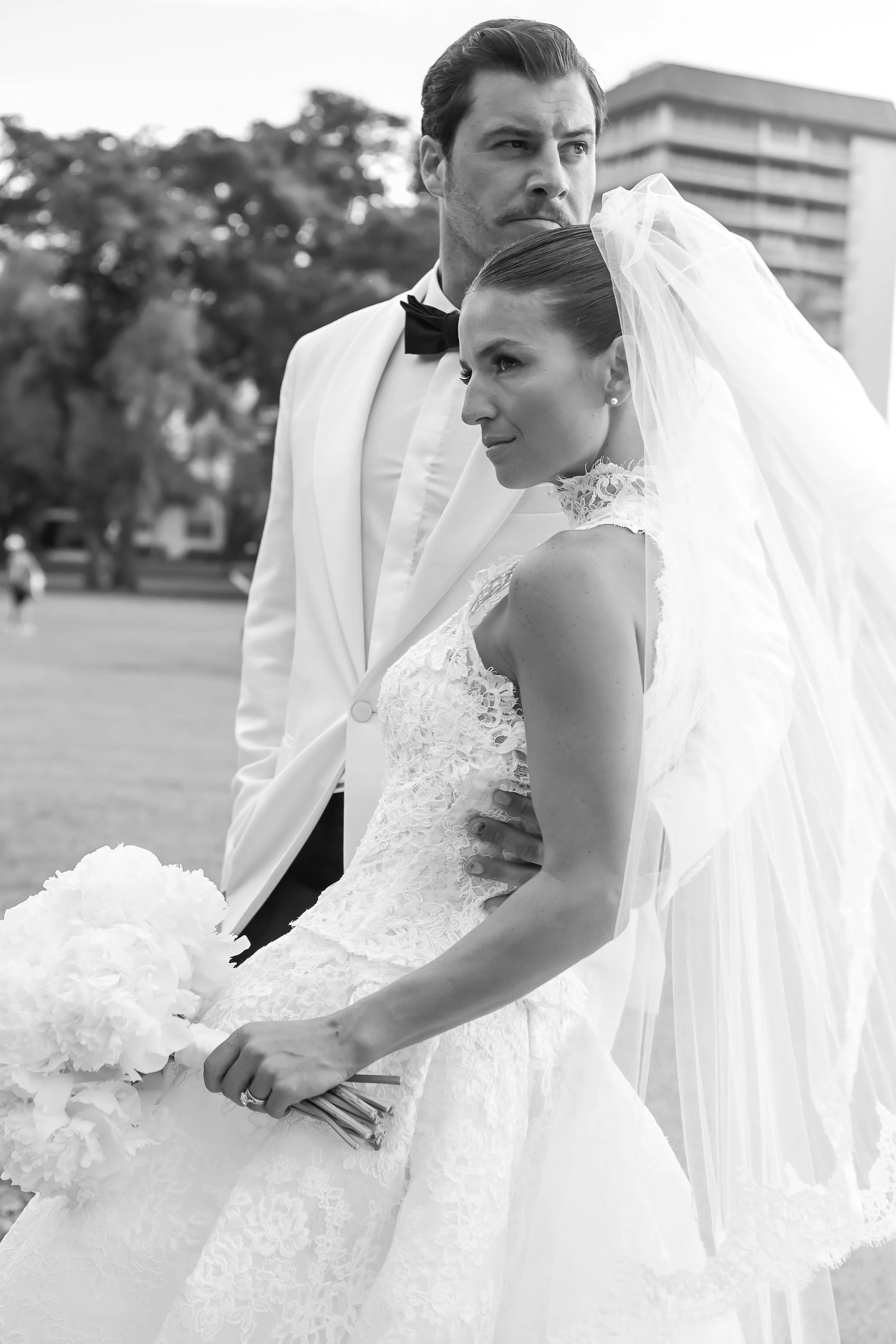 Black and white photo of a bride and groom outdoors, with the bride holding a bouquet and wearing a lace wedding gown and veil, and the groom in a tuxedo with a bow tie, standing close with a city building and trees in the background.