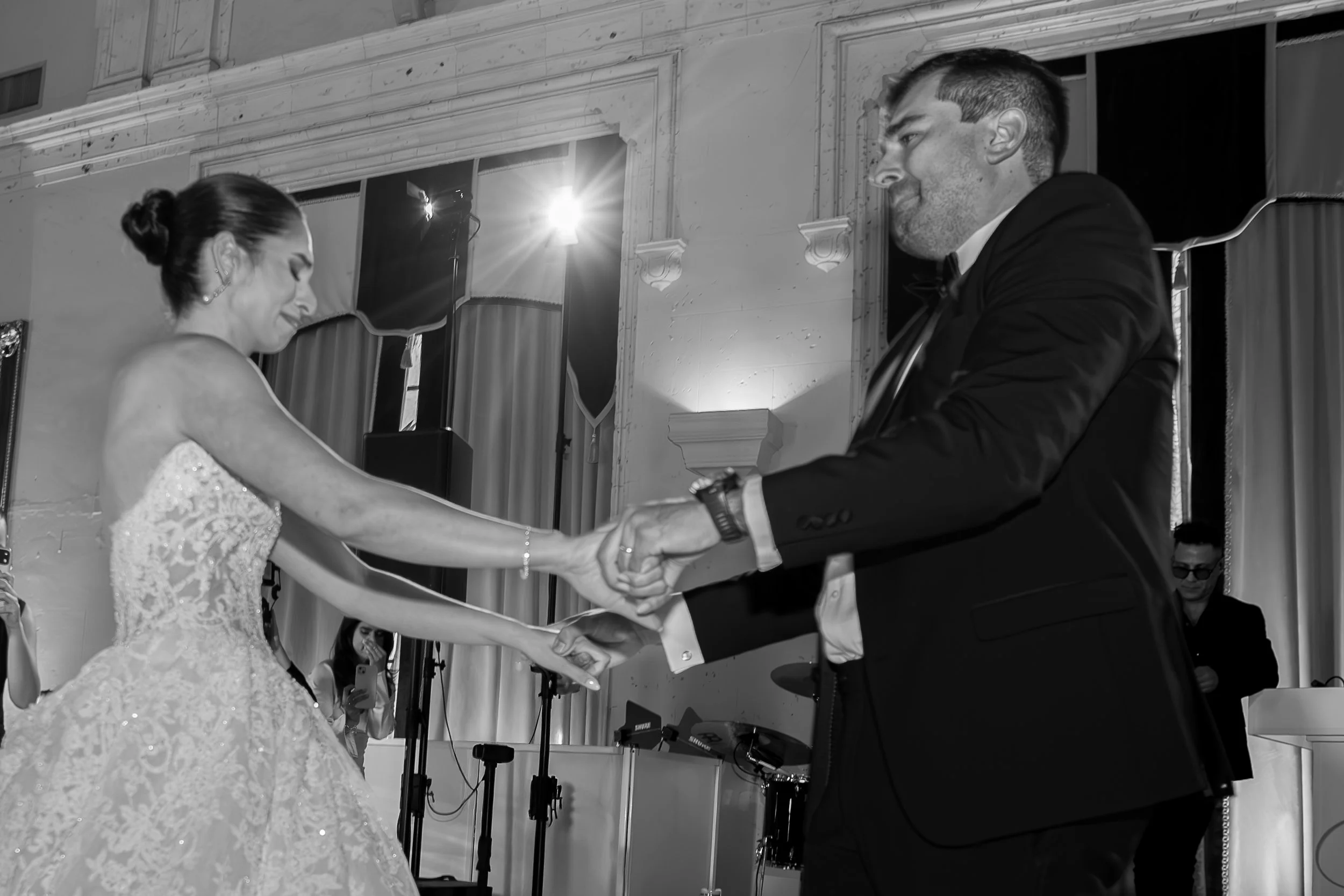 A bride and groom are holding hands during their wedding dance in a decorated ballroom. The bride is wearing a lace wedding gown, and the groom is dressed in a dark tuxedo with a bow tie.