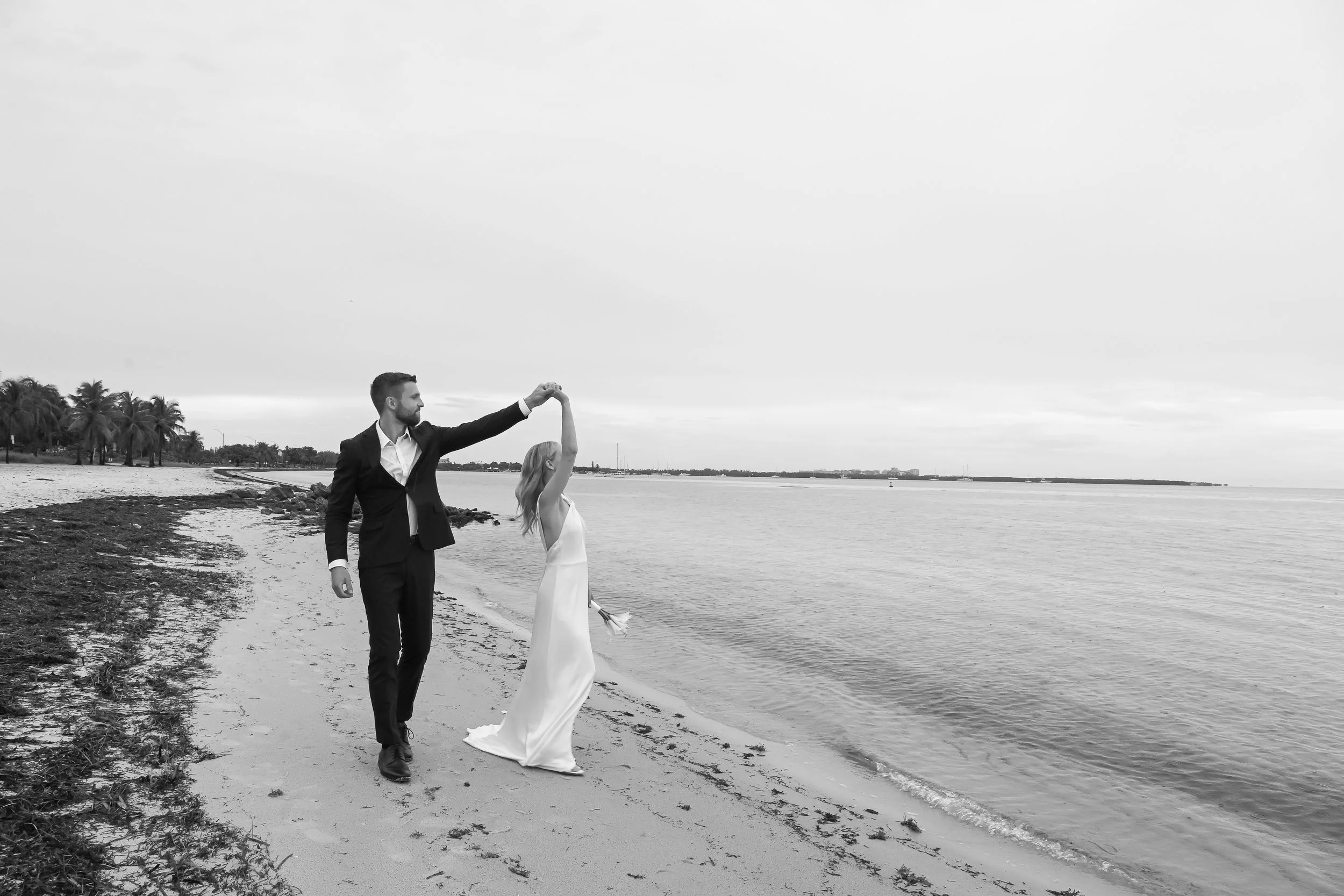 A black and white photo of a bride and groom on a beach, with the groom holding the bride's hand as they walk near the water.