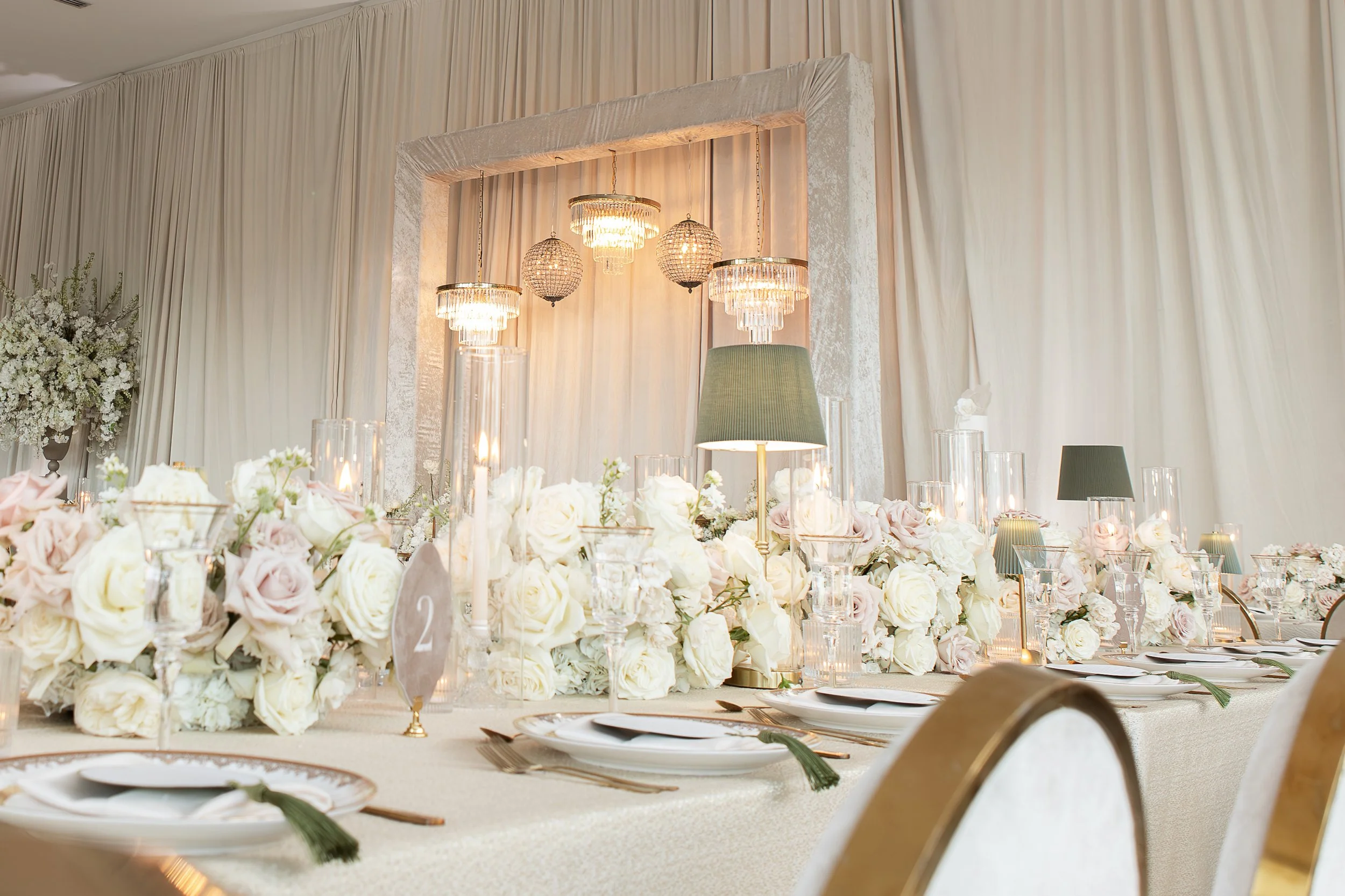 Elegant banquet table decorated with white and blush roses, candles, and glassware, set in a room with cream-colored curtains and a chandelier hanging above.