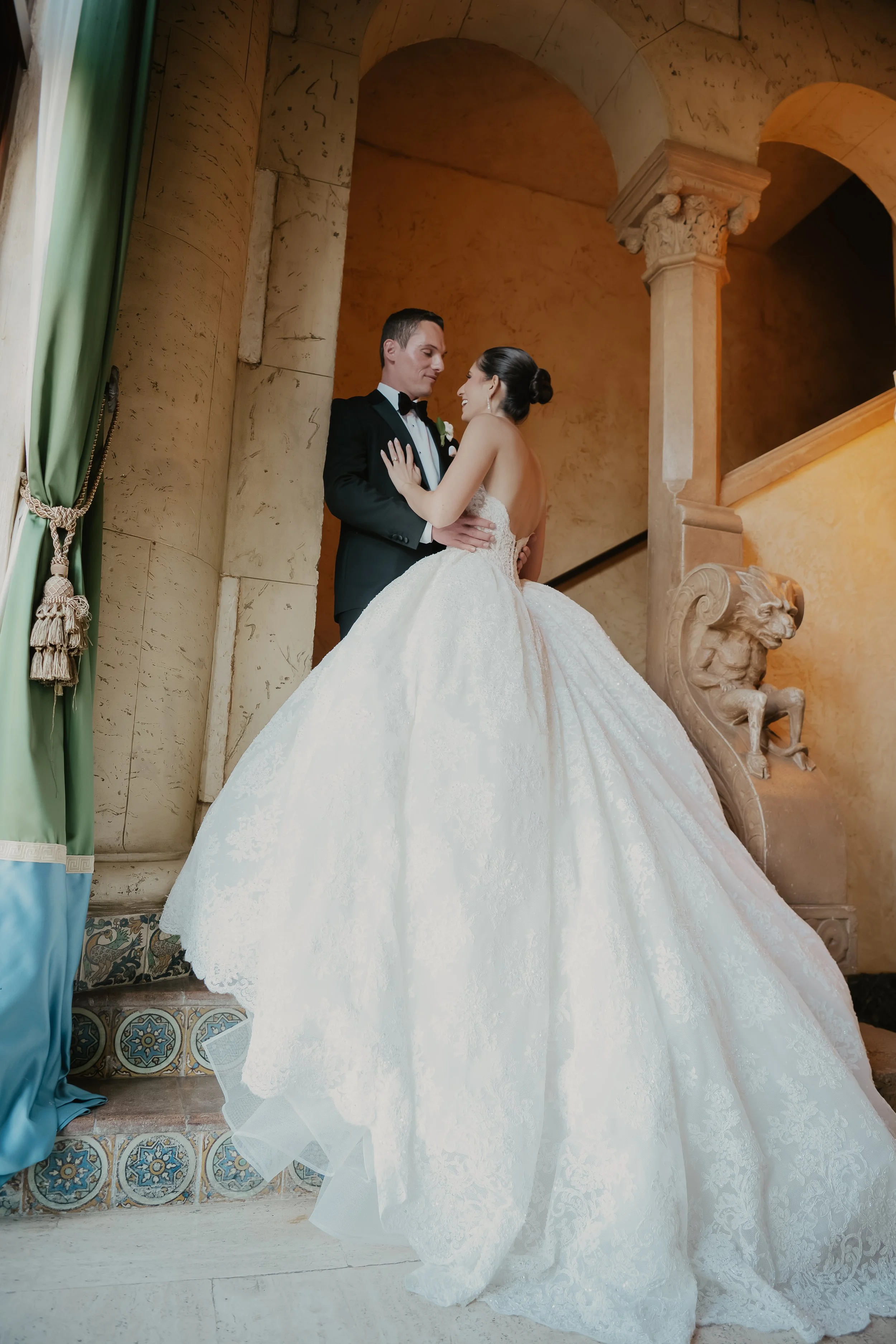 A bride and groom sharing a moment in an elegant, ornate indoor setting. The bride's white lace wedding gown has a full skirt, and the groom is dressed in a black tuxedo with a white shirt and black bow tie. They are smiling and looking at each other