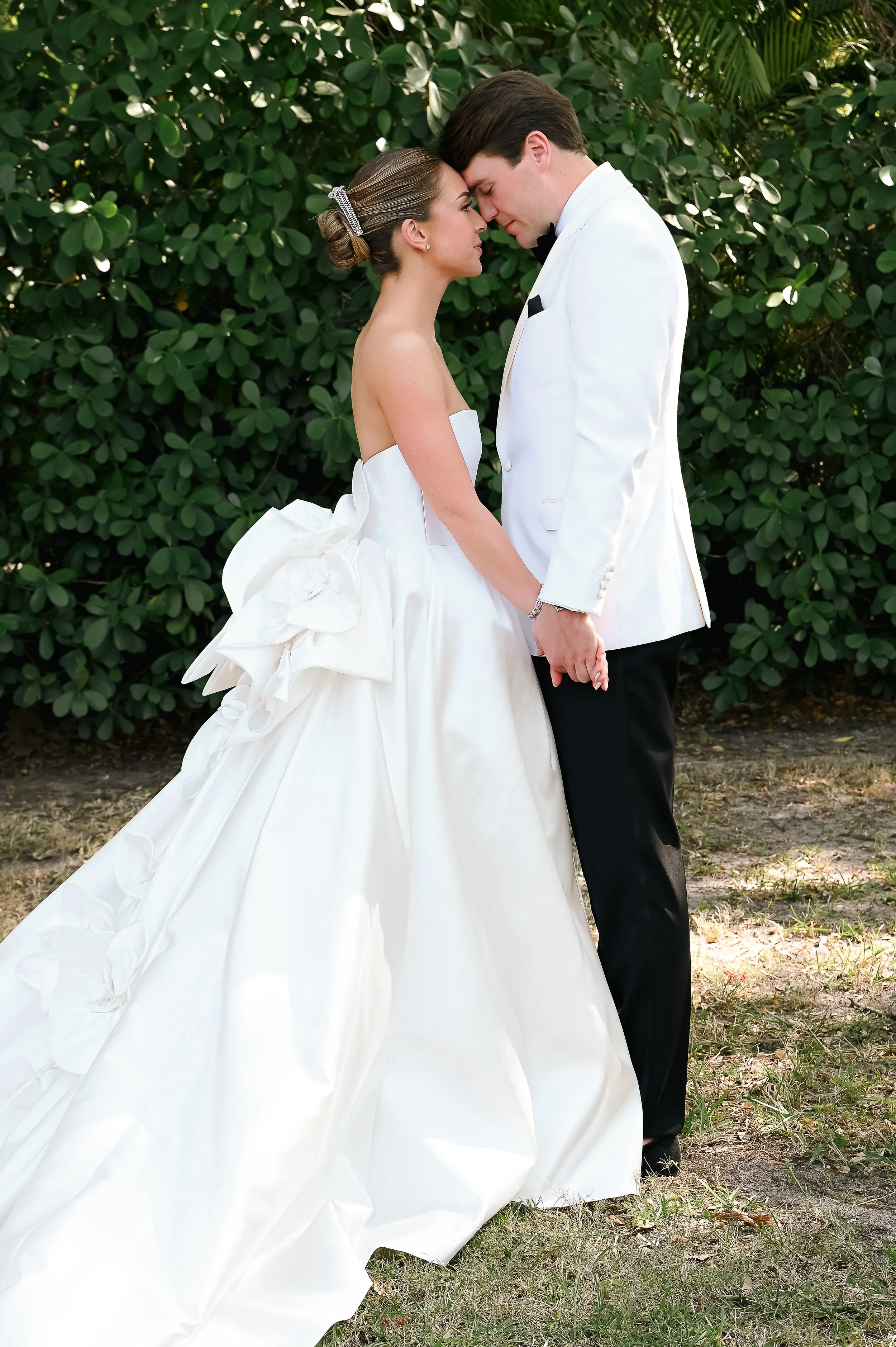 A bride and groom standing close with their foreheads touching outdoors, surrounded by green bushes. The bride is wearing a white strapless wedding gown with ruffle details, and the groom is dressed in a white tuxedo jacket with a black bow tie and b