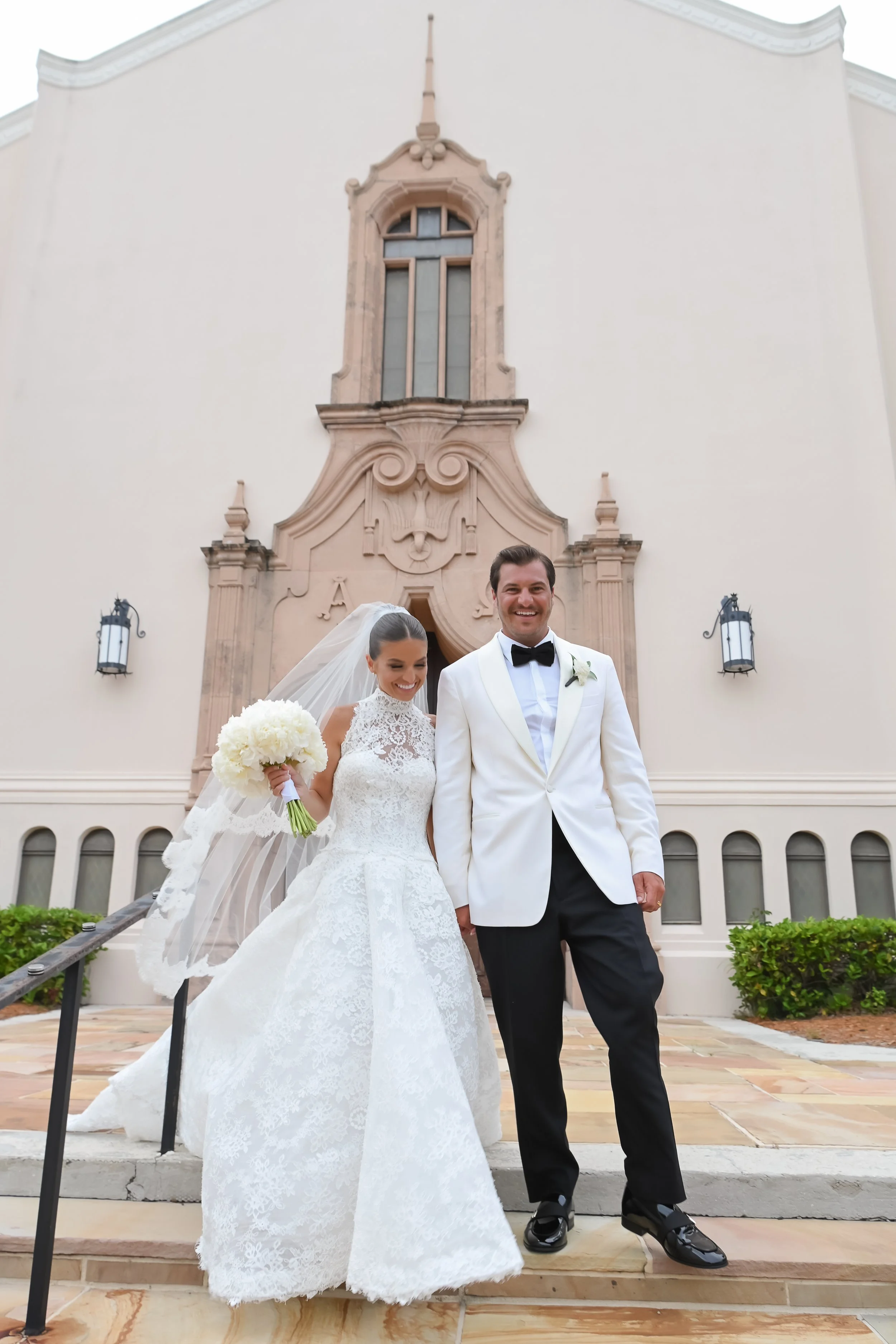 A bride and groom standing outside a white church building, smiling after their wedding. The bride wears a lace wedding gown and holds a bouquet of white flowers, while the groom wears a white tuxedo with a black bow tie. The background includes arch