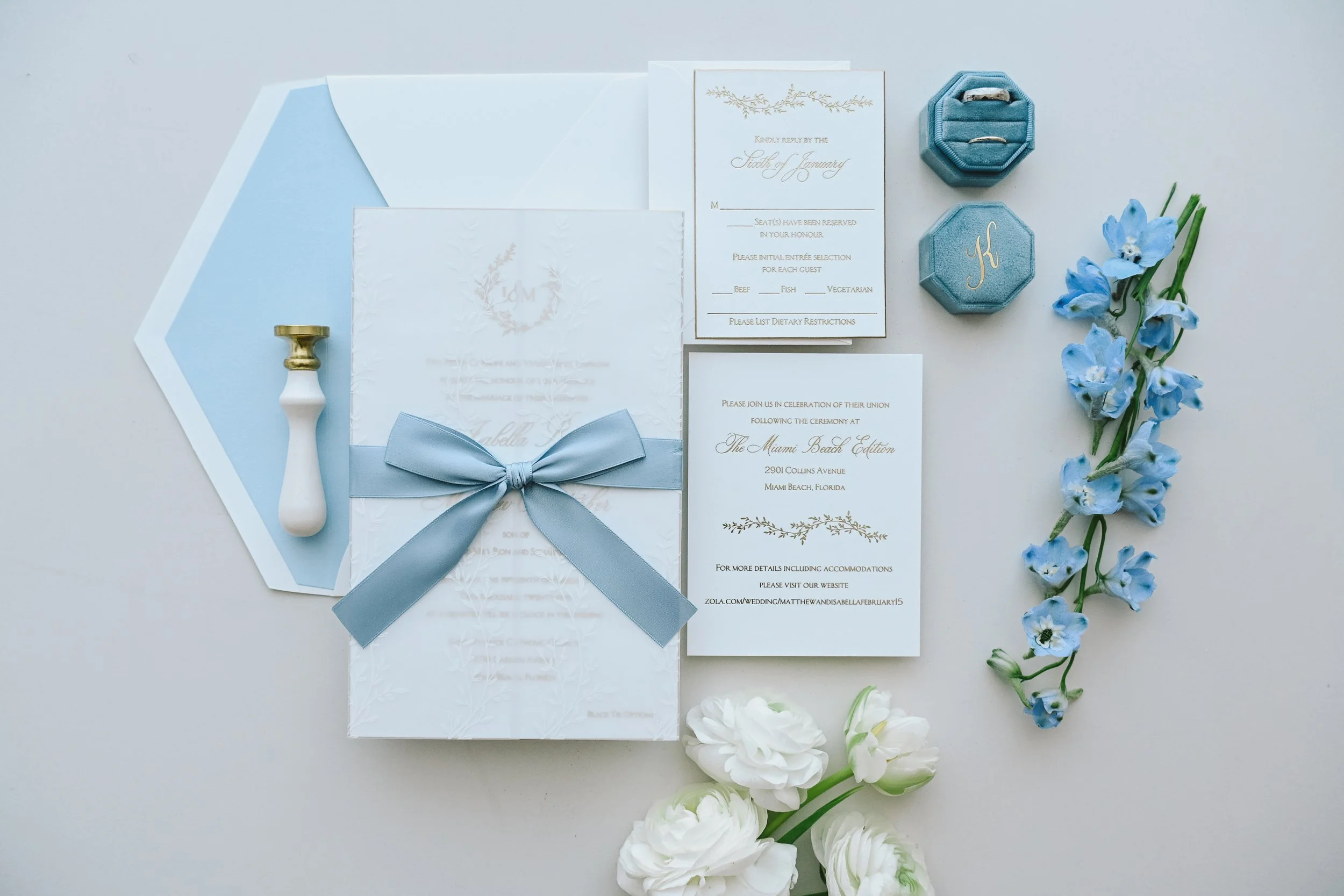 Wedding invitation suite with light blue accents, including invitation cards, a white envelope, a white and gold seal stamp, and light blue and white flowers on a white background.