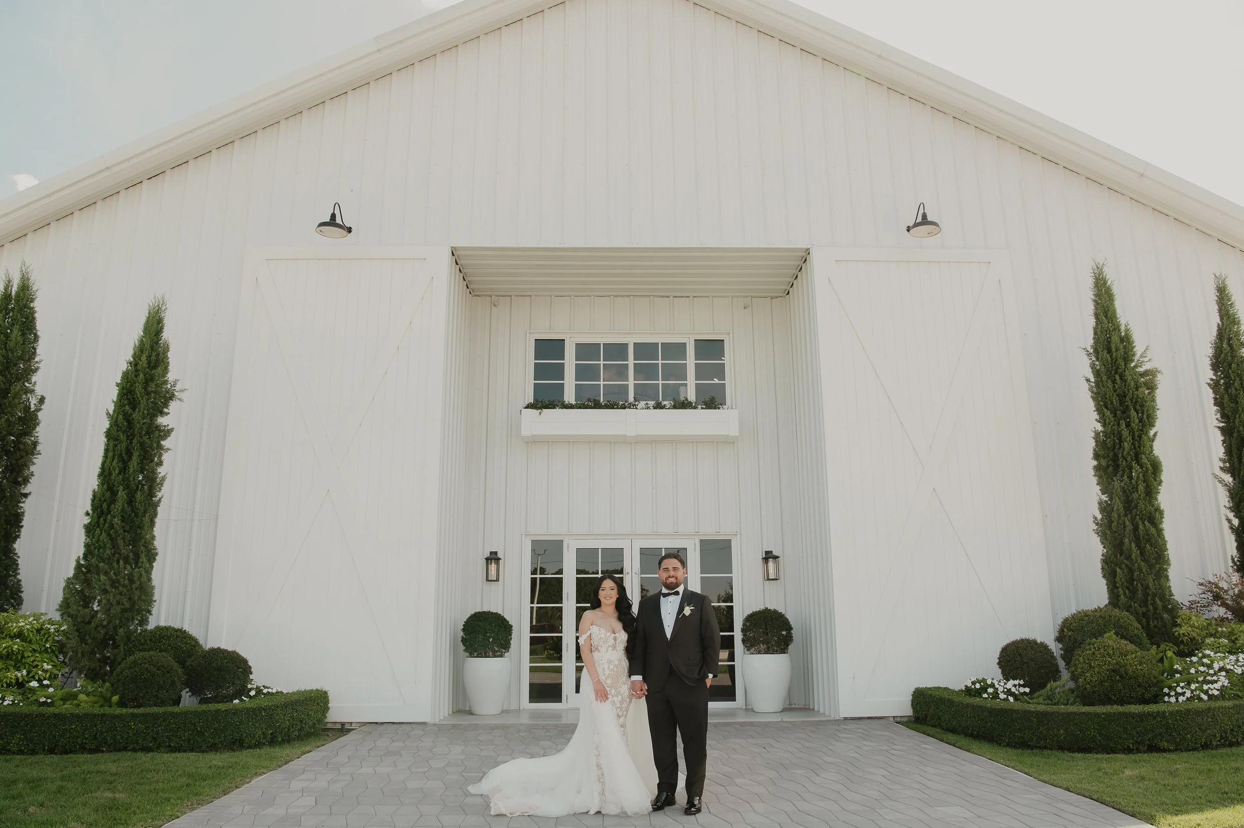 A bride and groom standing in front of a white barn with tall trees and manicured bushes on either side, during daytime.
