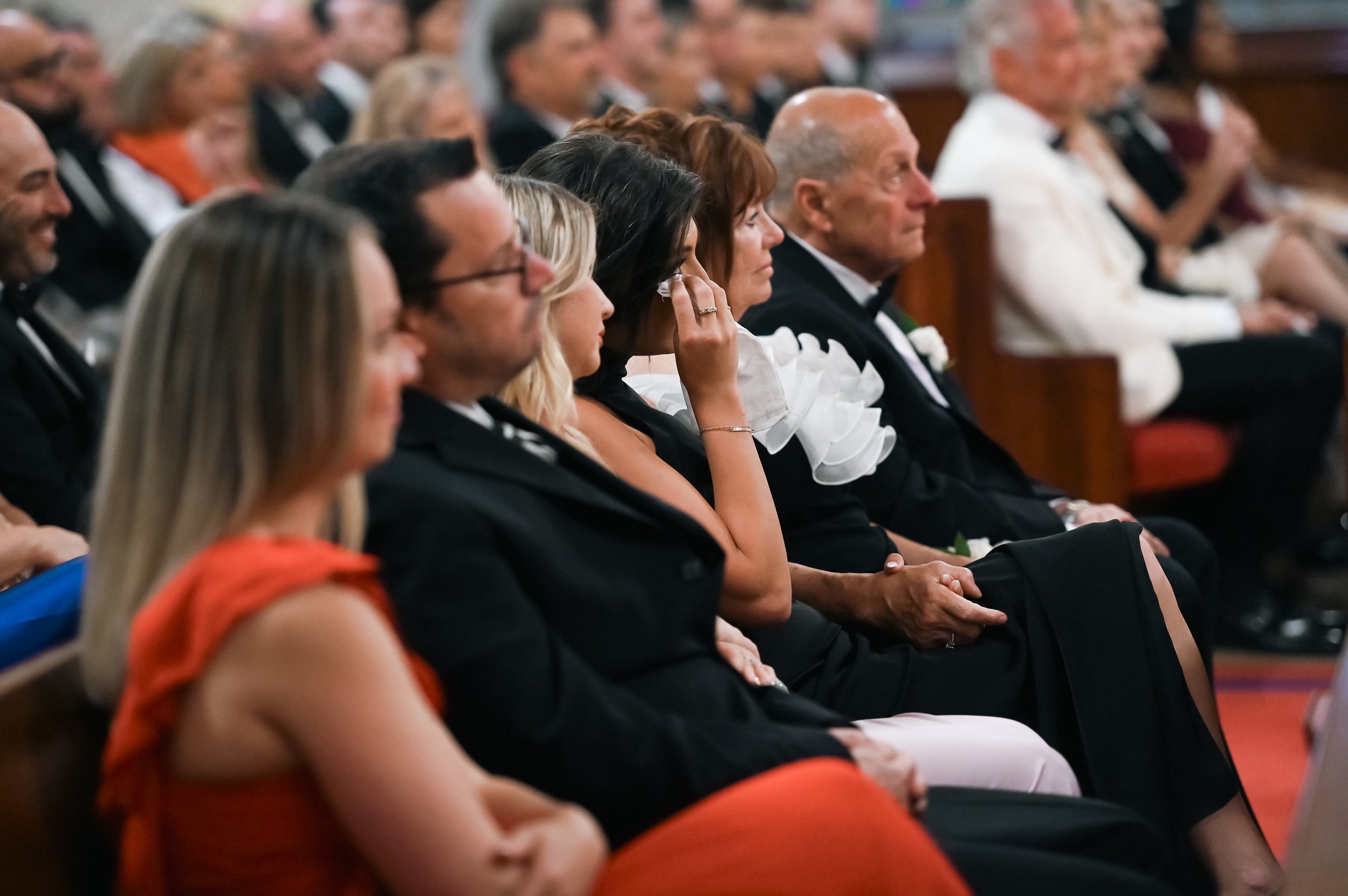 People in formal attire sitting in an auditorium, attending an event or ceremony, with women wearing dresses and men in tuxedos or suits.