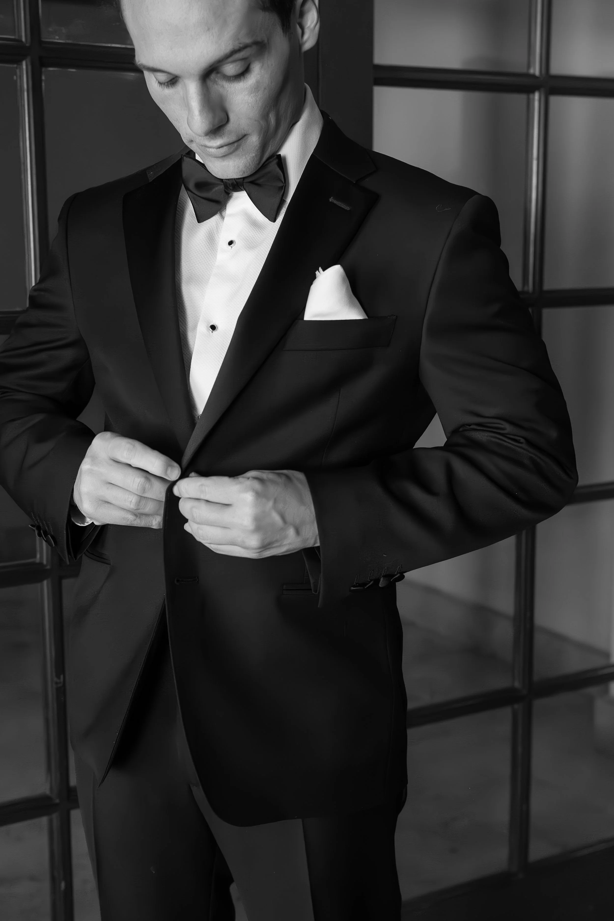 A man dressed in a black tuxedo with a bow tie and white shirt, adjusting his jacket, standing in front of a dark wall with framed sections.