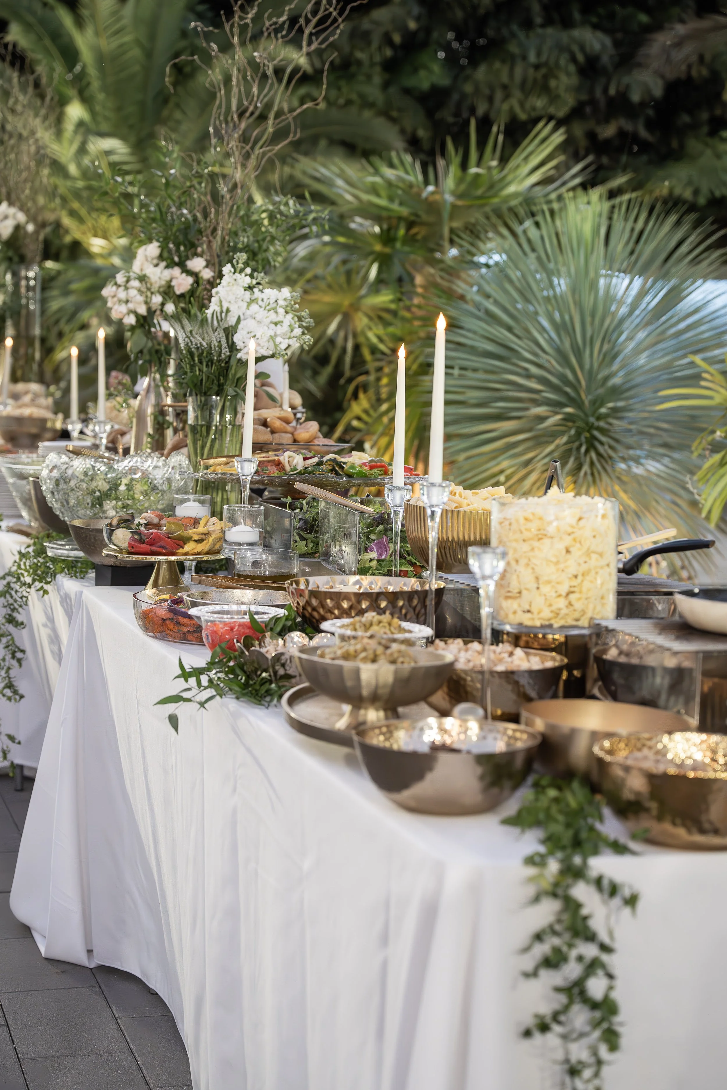 A long buffet table draped in a white tablecloth, decorated with candles, glassware, and an assortment of food dishes, set outdoors against a background of lush green tropical plants and foliage.
