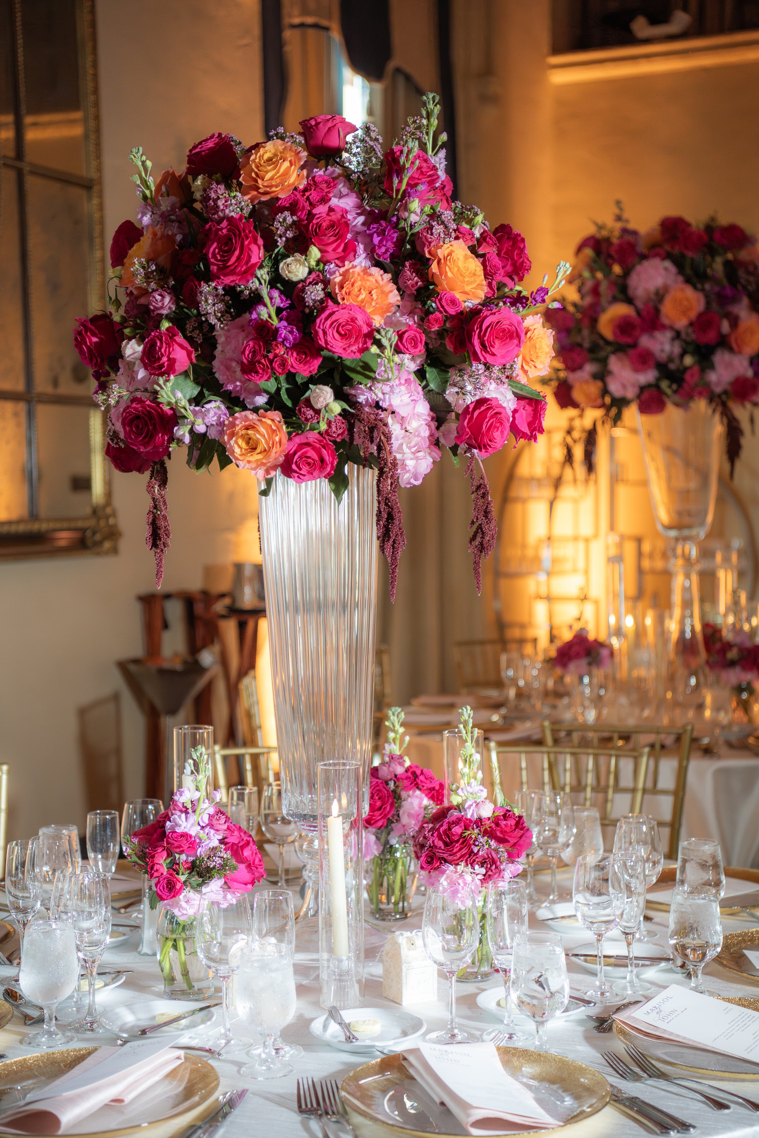 A tall clear glass vase filled with a large, colorful arrangement of pink, orange, and purple flowers, with some greenery, set on a banquet table at an elegant event venue with candle lighting and gold chairs.