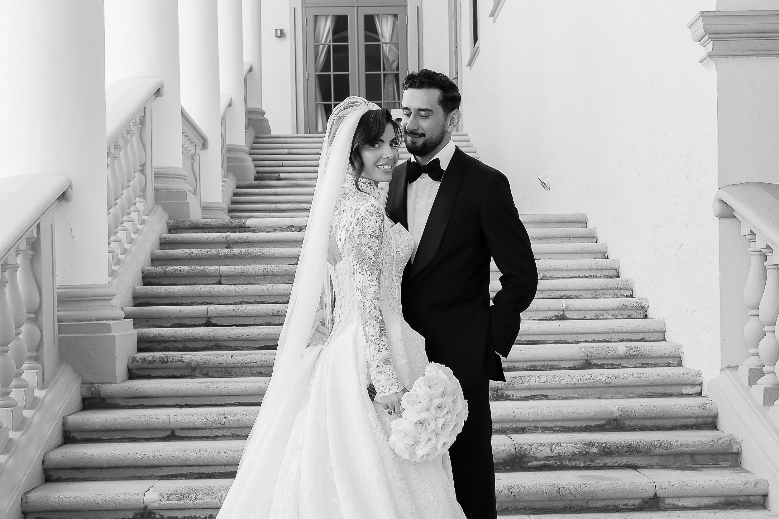 A bride and groom standing on a staircase outside a building at night, smiling and looking at each other. The bride holds a bouquet of roses, wearing a lace wedding dress and veil. The groom is dressed in a tuxedo with a bow tie.