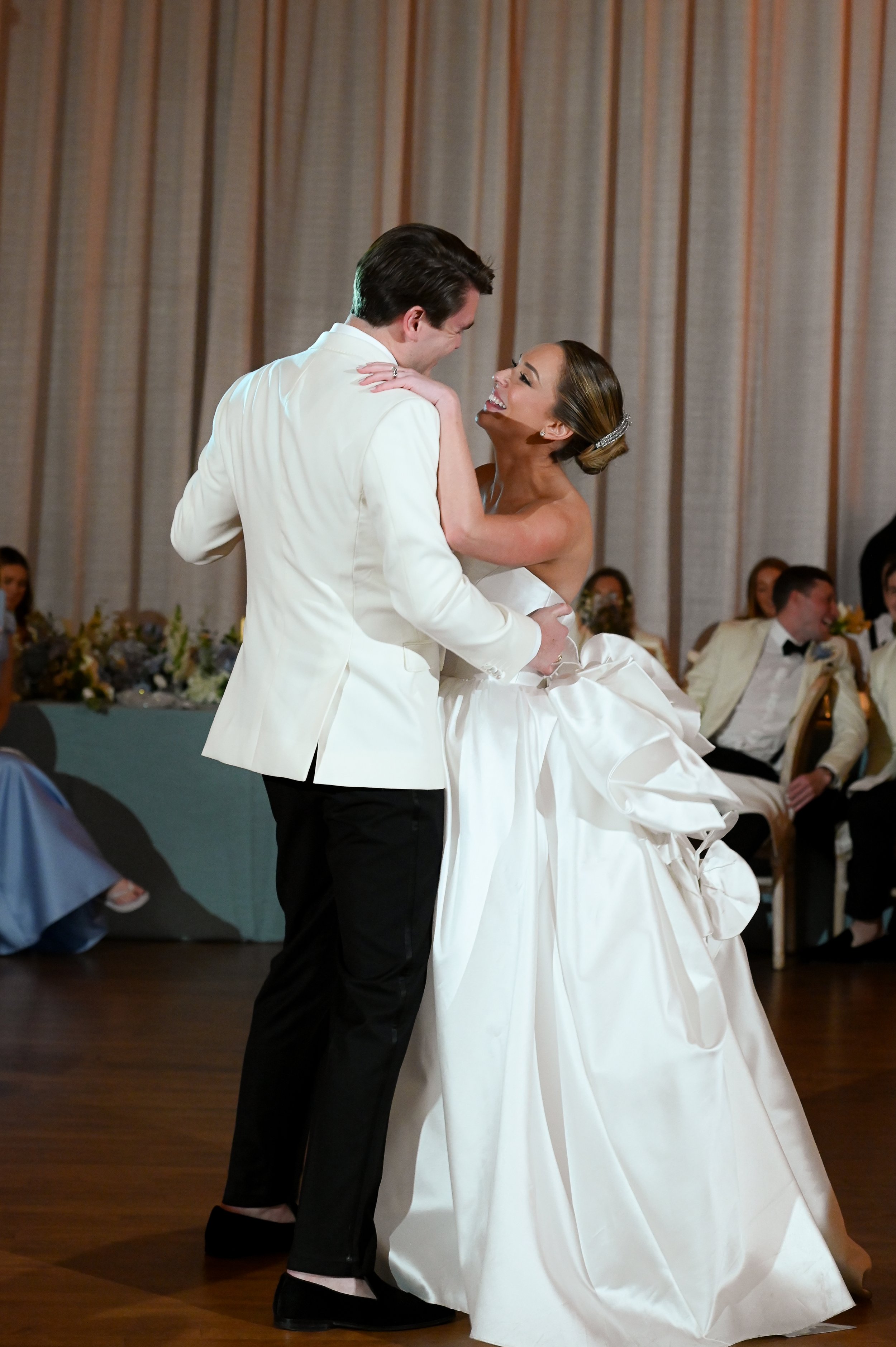 A bride and groom dancing at their wedding reception. The groom is wearing a white tuxedo jacket and black pants, and the bride is in a strapless white gown with an updo hairstyle and a headband. They are smiling and looking at each other.