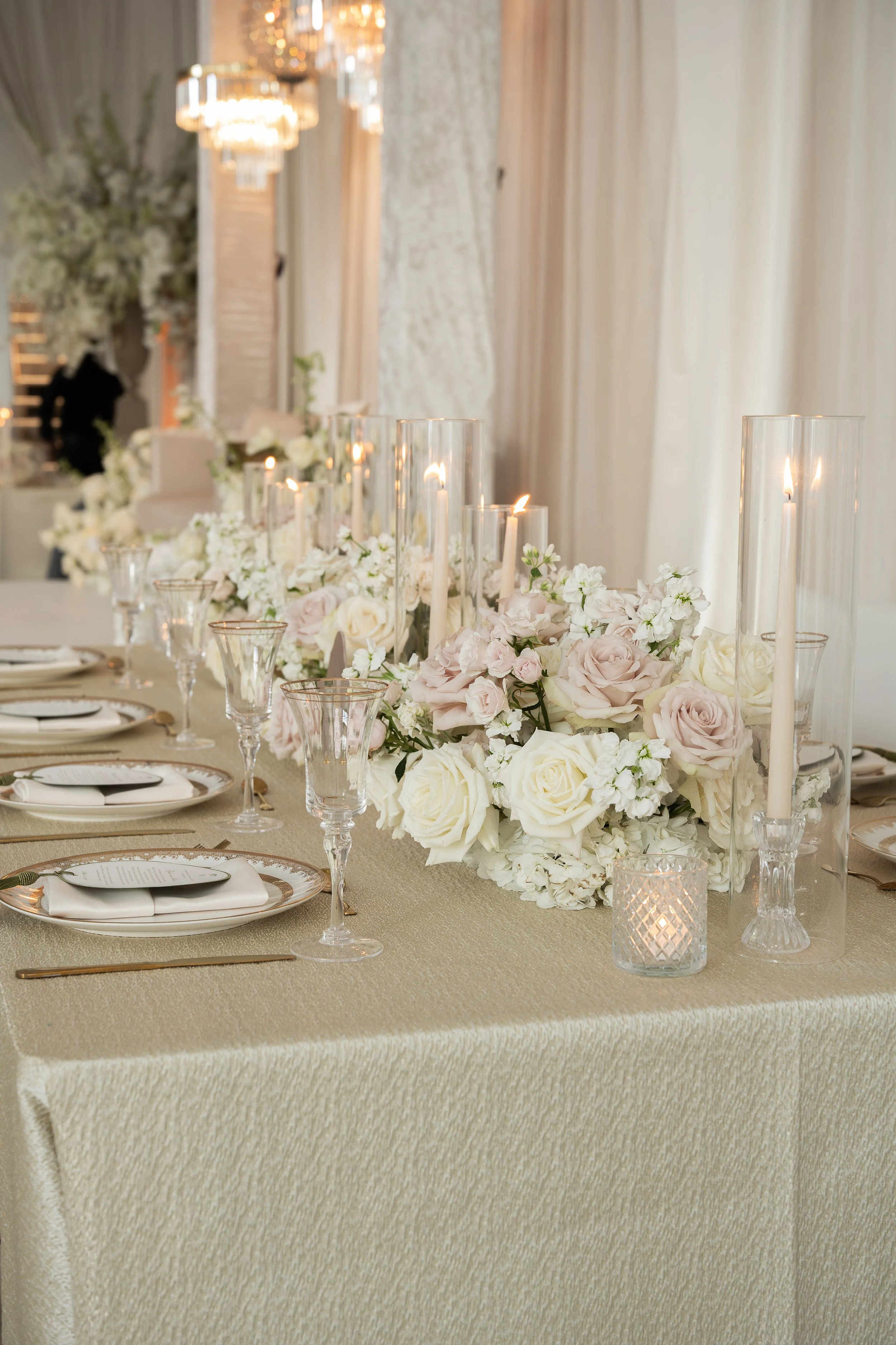 Elegant banquet table with cream-colored tablecloth, white roses, blush pink flowers, lit candles in tall glass holders, and small votive candles, set in a decorated room with chandeliers and drapes.