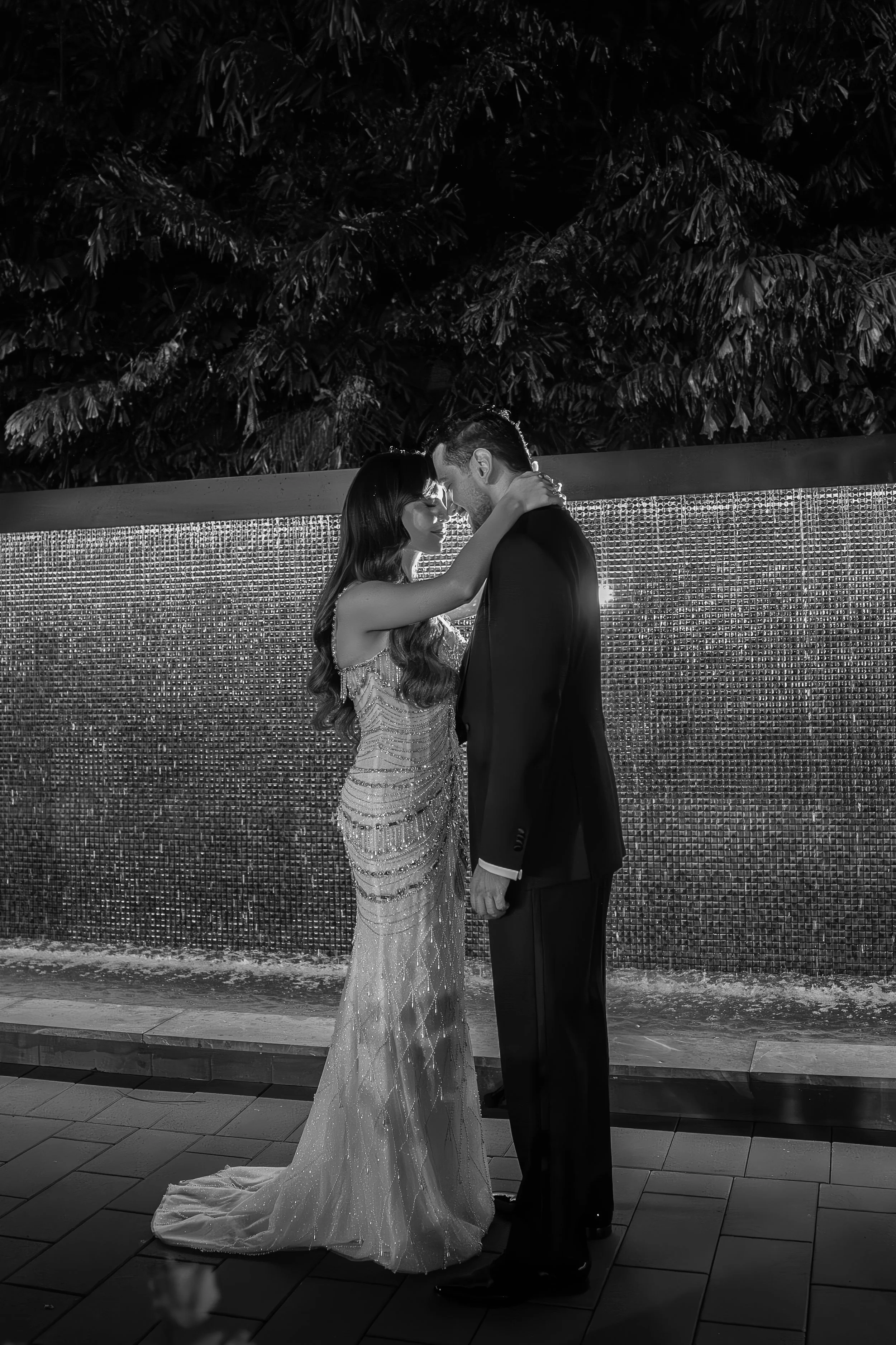 A couple dressed in formal attire, standing close with foreheads touching and arms around each other, in front of a textured metallic wall, outdoors at night.