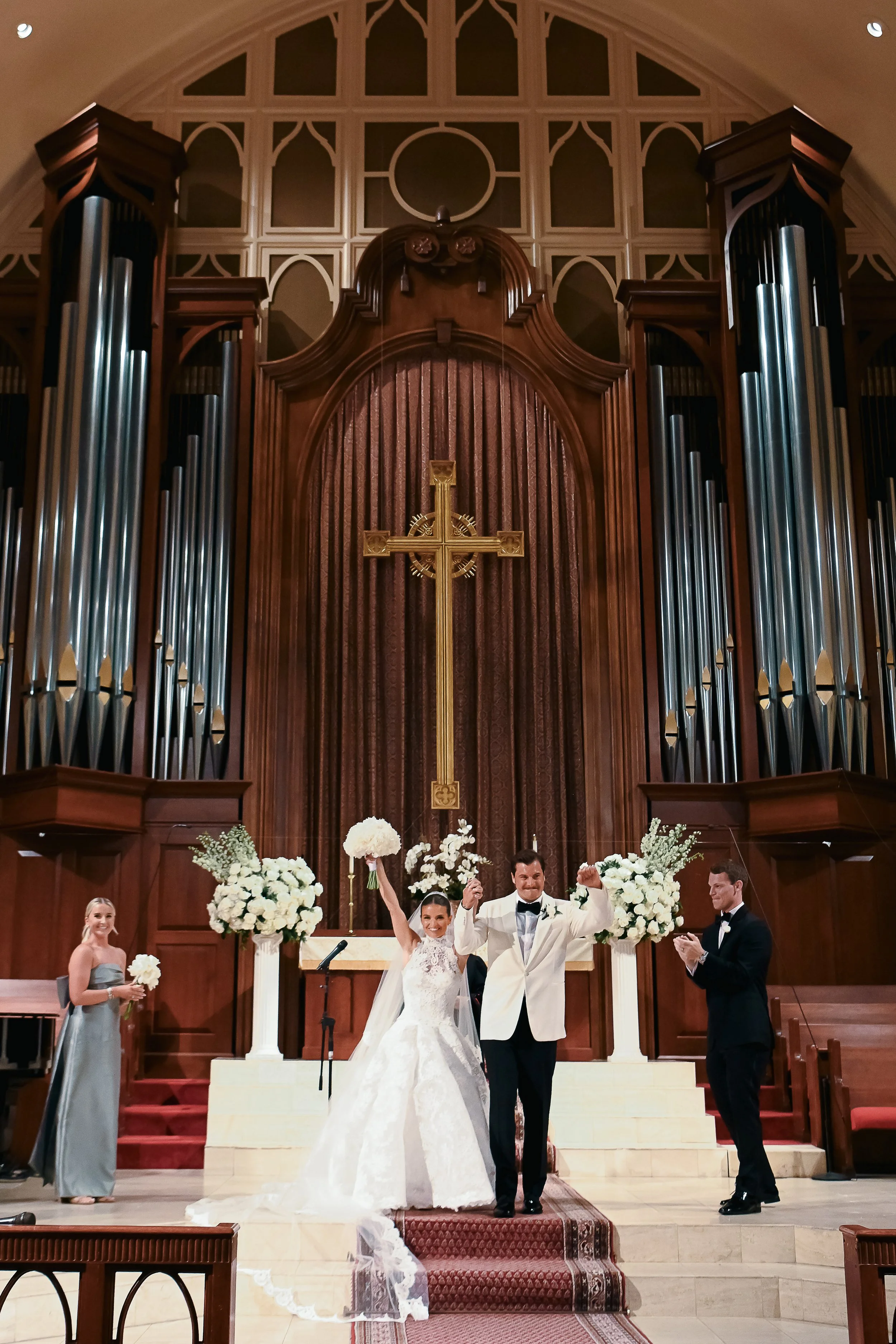 A bride and groom celebrating at their wedding ceremony in a church, standing on the steps of the altar with flowers around them, while the bridesmaid and groomsman stand to the sides.
