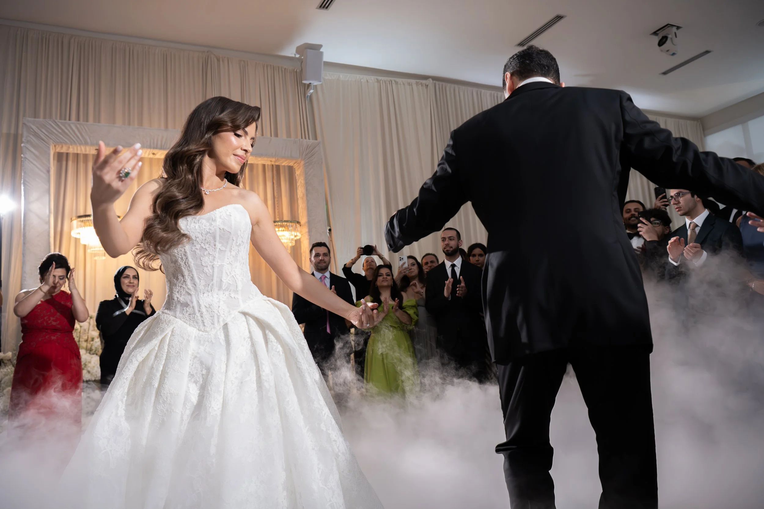 A bride in a white wedding dress and a groom in a black tuxedo dancing at a wedding reception surrounded by guests.