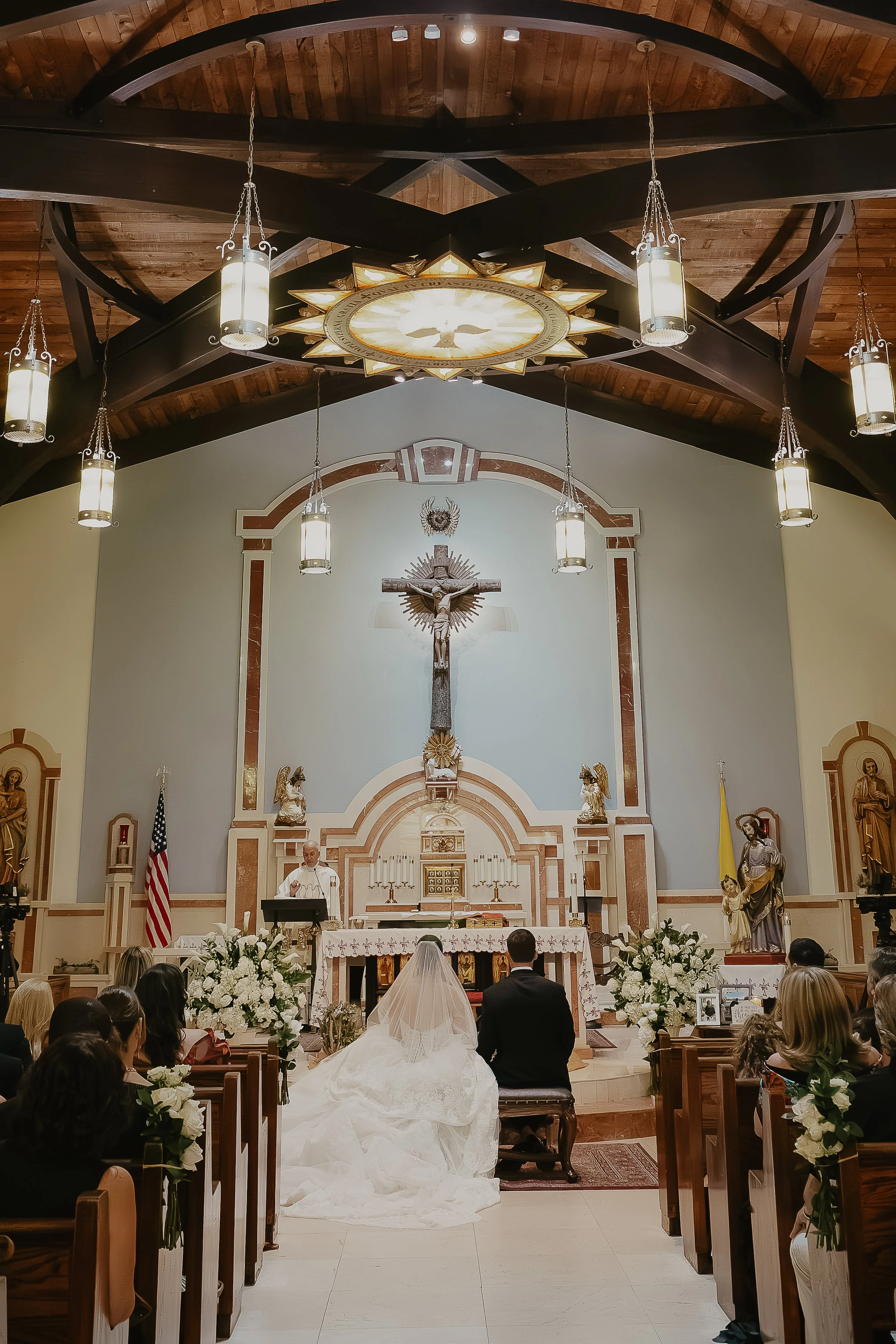 Inside a church, the bride and groom are kneeling at the altar during a wedding ceremony. The bride is wearing a white wedding dress with a veil, and the groom is dressed in a black suit. The altar features religious statues, a crucifix, and floral a