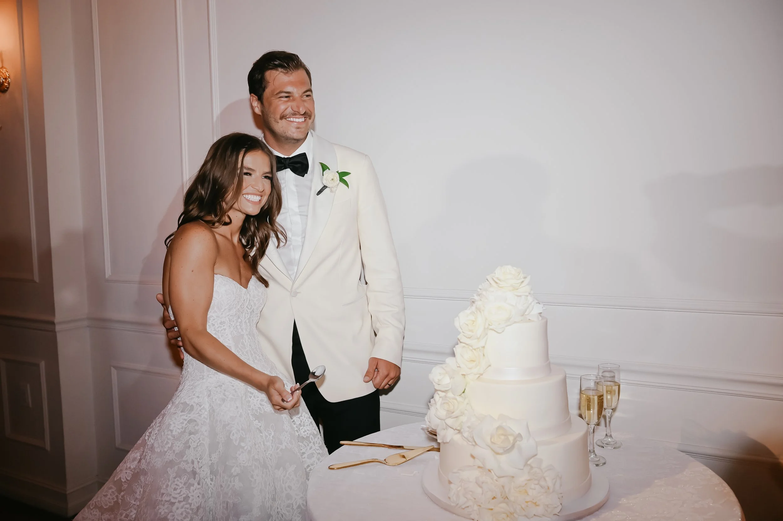 A bride and groom cut a wedding cake together at their wedding reception. The bride wears a white lace wedding dress, and the groom wears a white tuxedo with a black bow tie. The wedding cake is decorated with white roses and is placed on a table wit