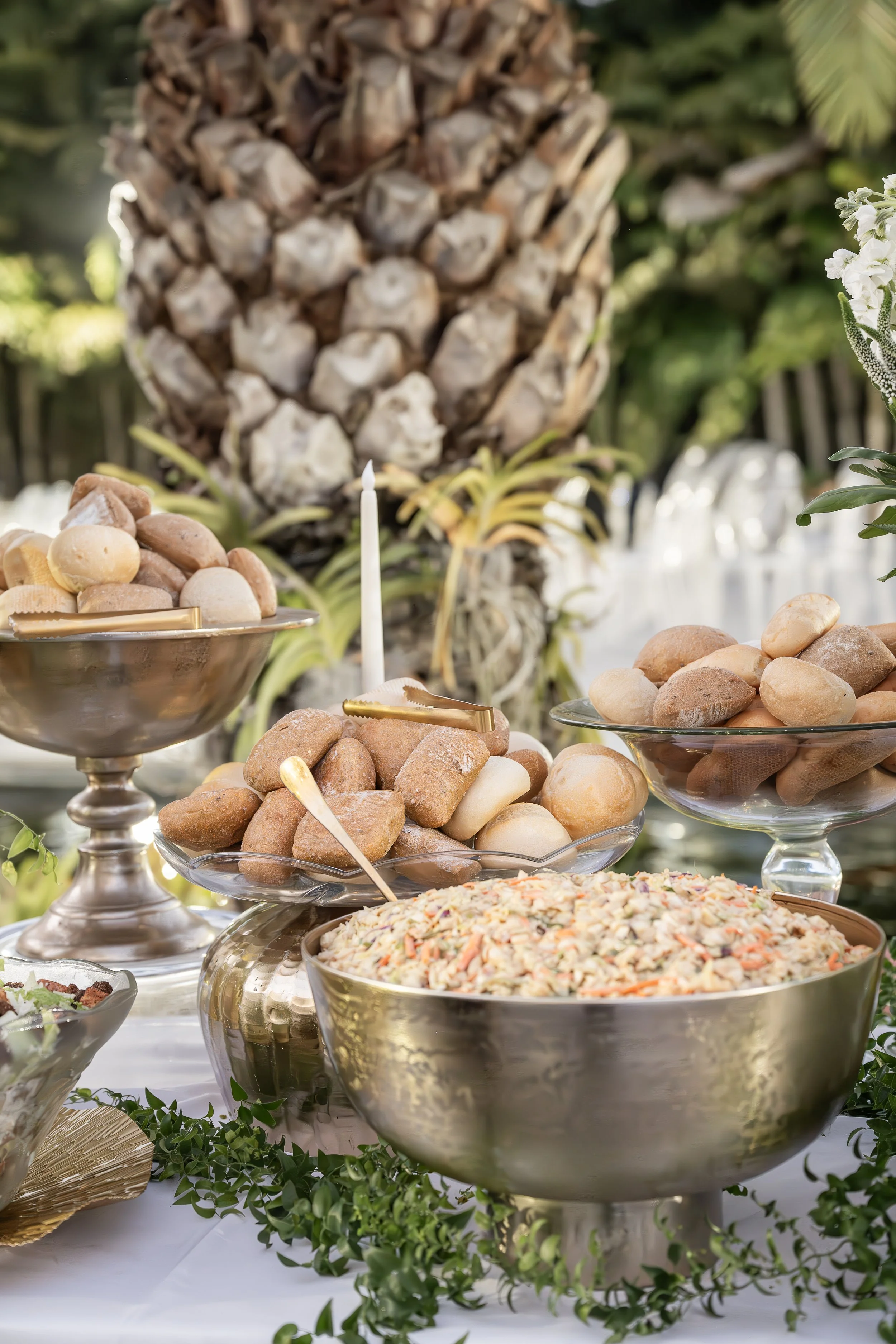 A variety of bowls filled with food such as potato salad, and a large pineapple in the background, with greenery and white flowers on a buffet table.