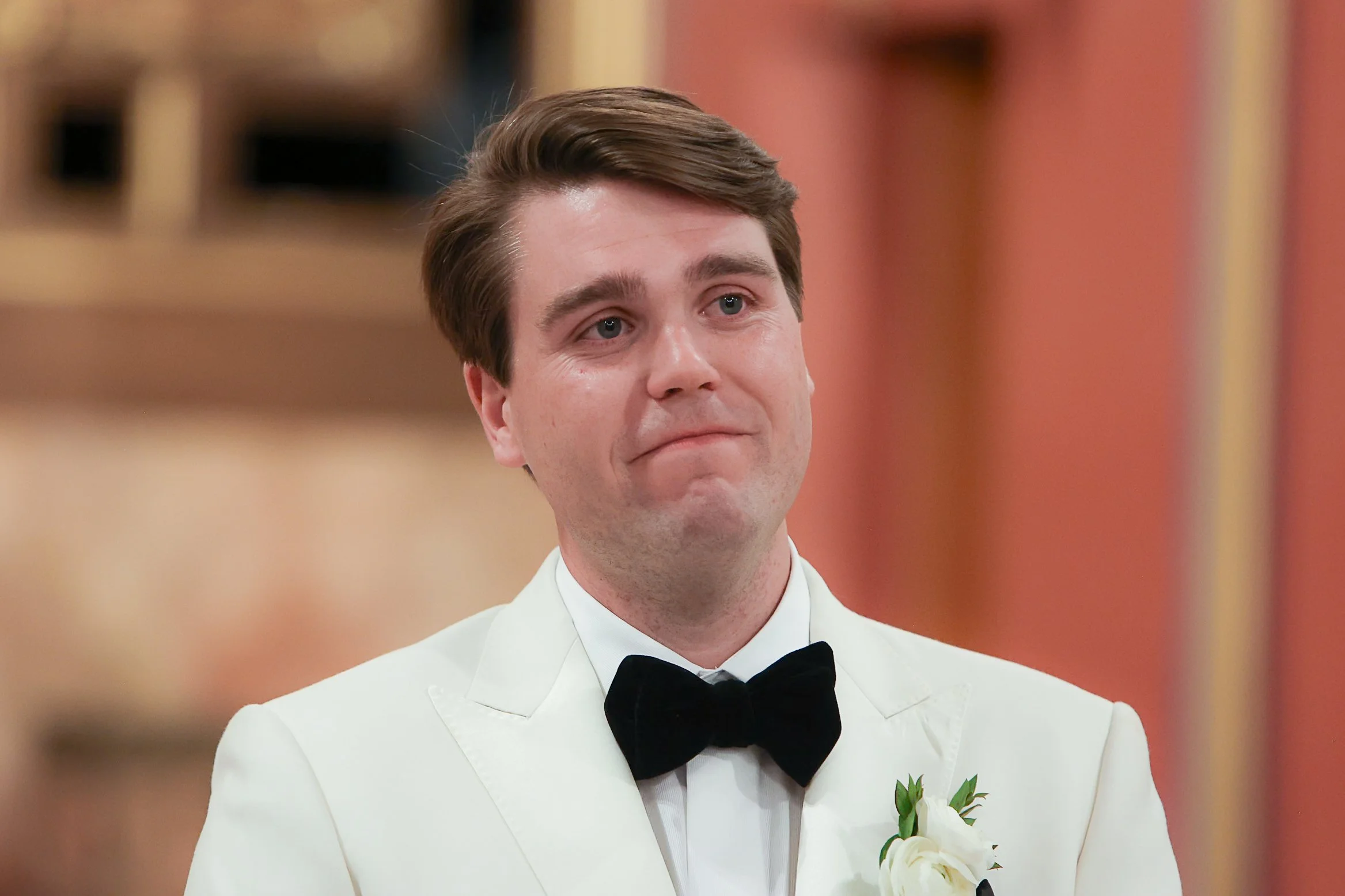 A man dressed in a formal white tuxedo with a black bow tie, standing indoors. He has light skin and brown hair, styled neatly. There is a small bouquet of white flowers with greenery pinned on his tuxedo's left lapel. The background is blurred, show