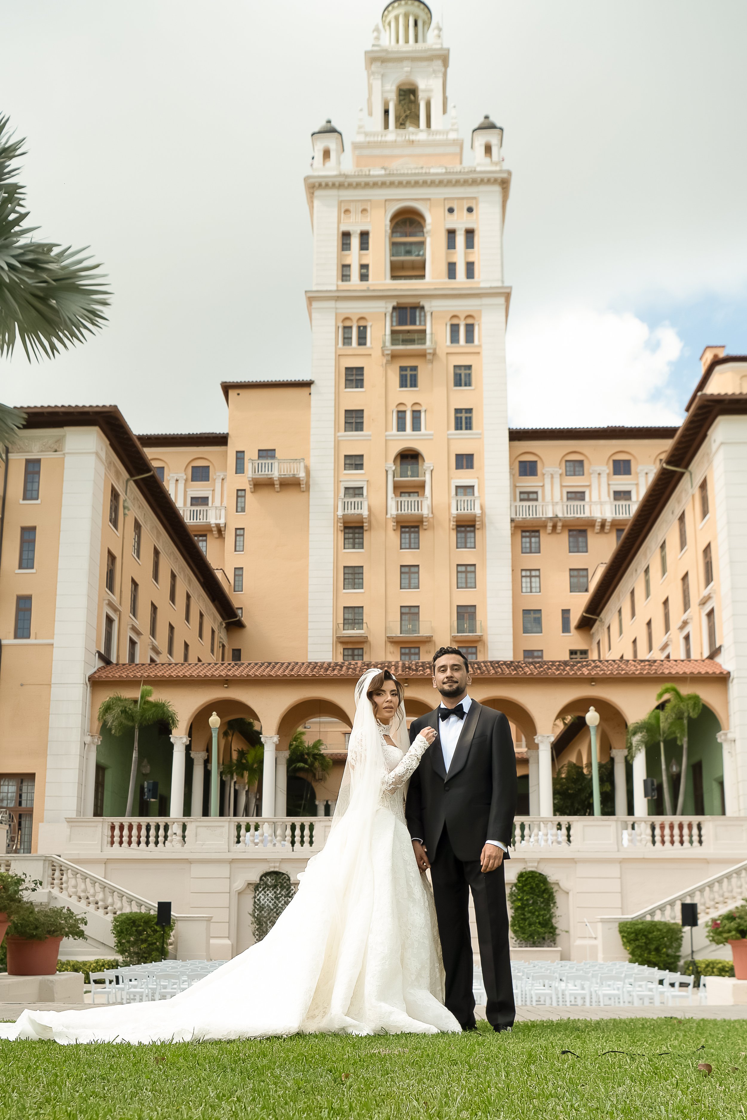 A wedding couple standing outdoors in front of a large, elegant hotel or building with a tower. The bride is in a white gown and veil, and the groom is in a black tuxedo. They are holding hands and looking at the camera. The setting is lush with gree