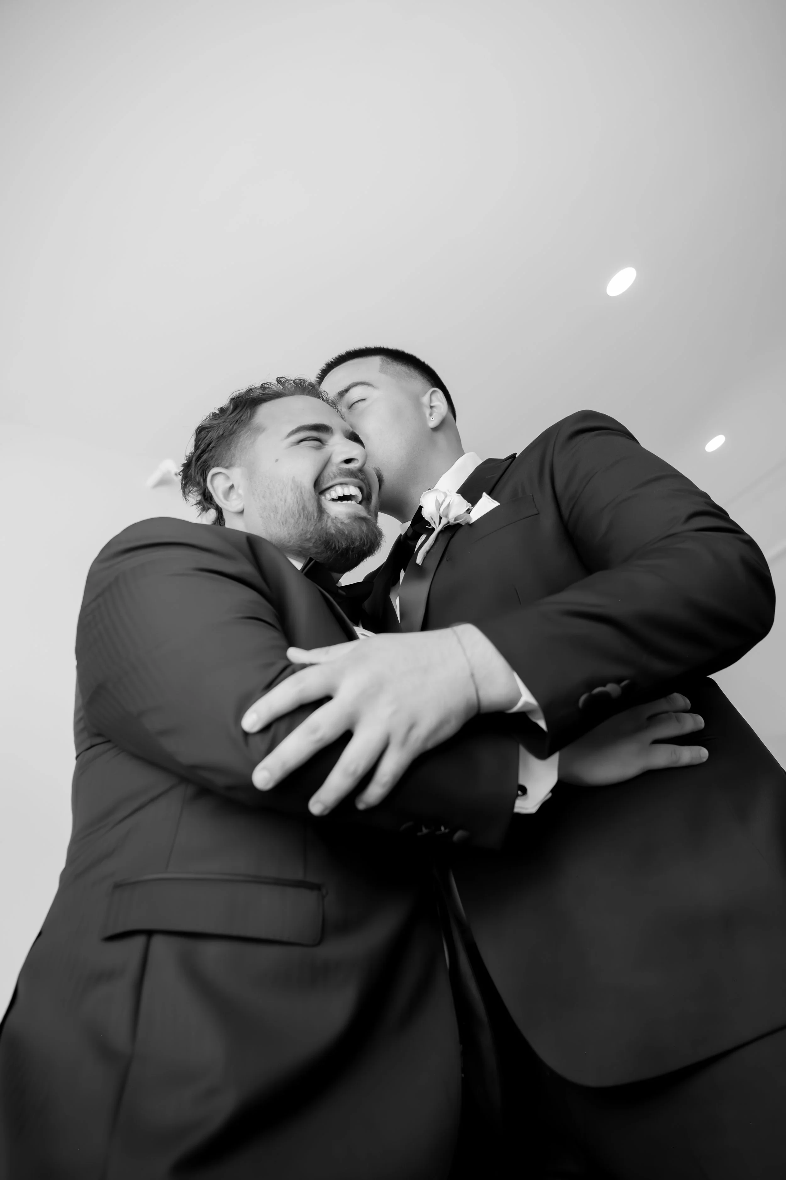 Two men in tuxedos embrace and kiss at a wedding celebration, with joyful expressions and close physical contact.
