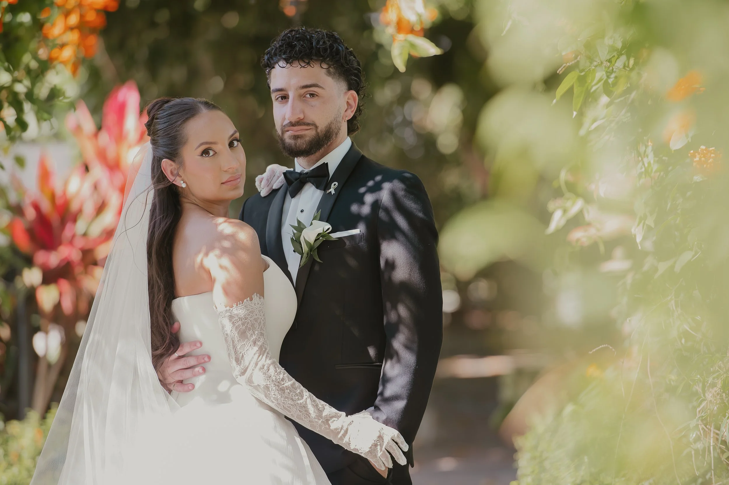 A bride and groom standing outdoors surrounded by greenery, with the bride in a white lace wedding gown and veil, and the groom in a black tuxedo with a bow tie, posing for a photo on their wedding day.