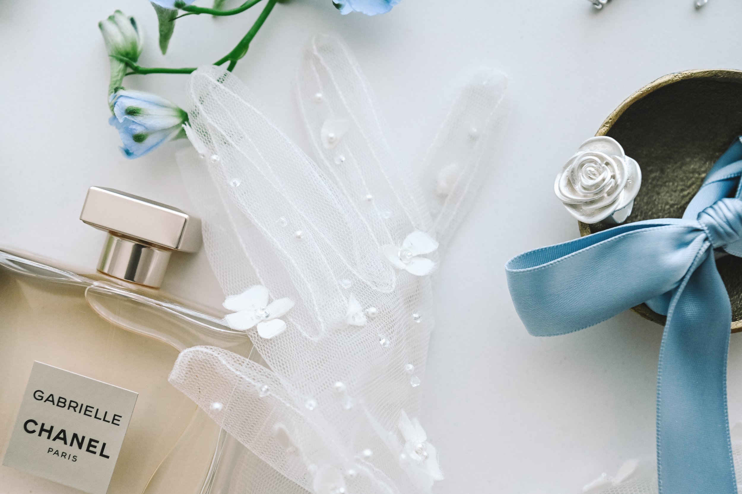 A collection of accessories including a white Chanel perfume bottle, a pair of angel wing earrings with pearl accents, a blue ribbon hair bow, and a white decorative rose, displayed on a white surface.