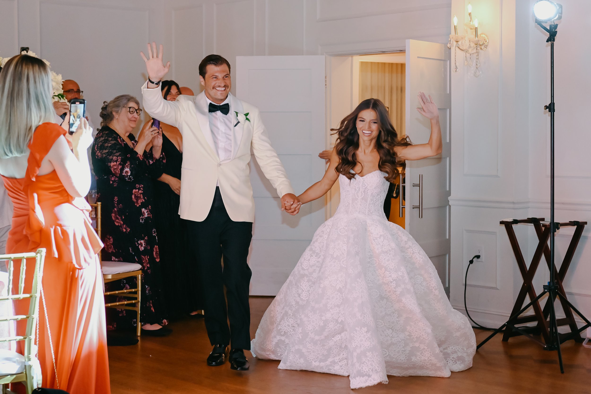 A wedding reception scene with a bride and groom holding hands and waving, surrounded by guests, with a bright light and decor in the background.