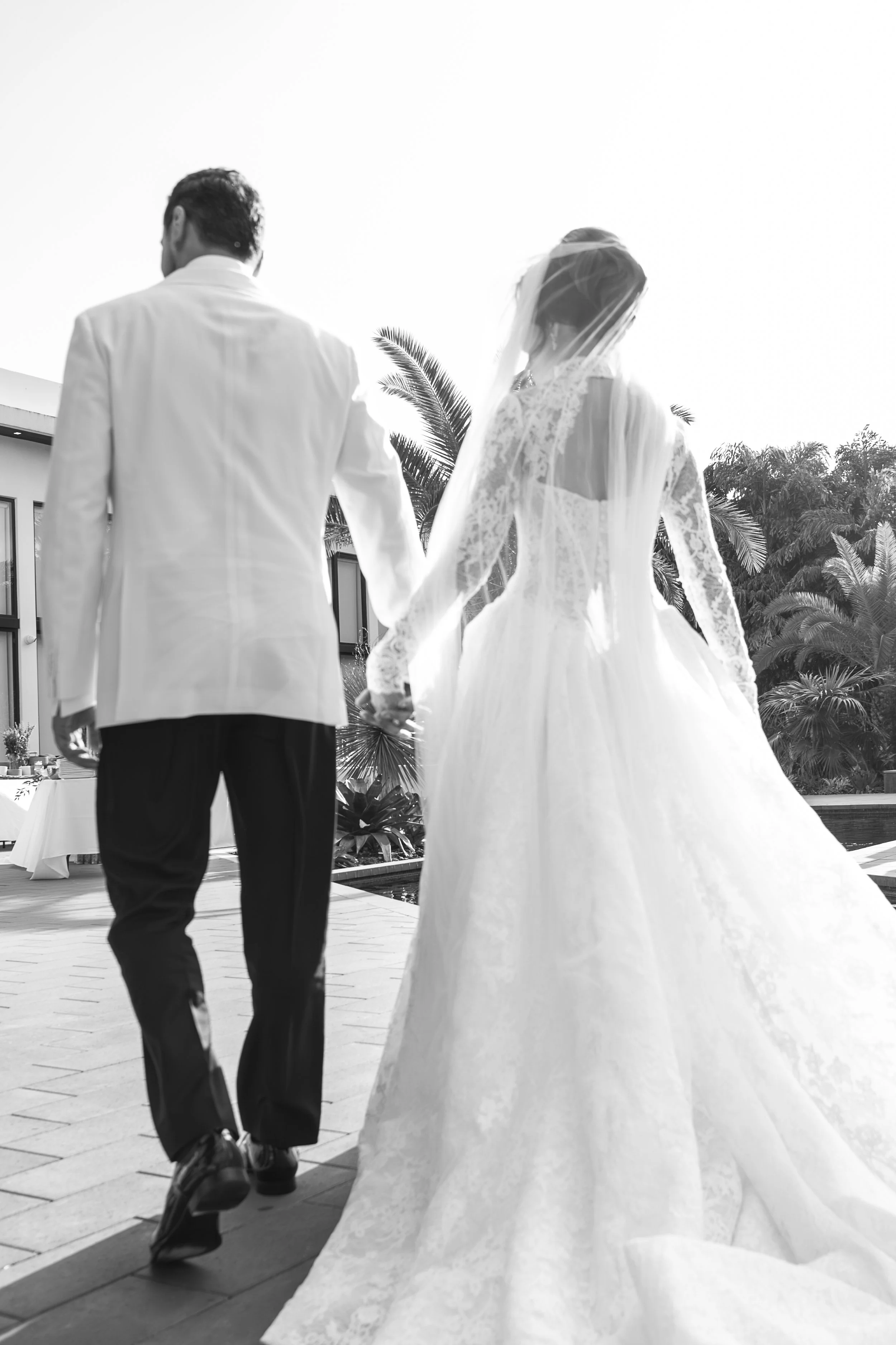 A black-and-white photograph of a bride and groom holding hands and walking outdoors. The bride is wearing a lace wedding gown with a veil, and the groom is dressed in a white jacket and dark pants. There are palm trees and a building in the backgrou