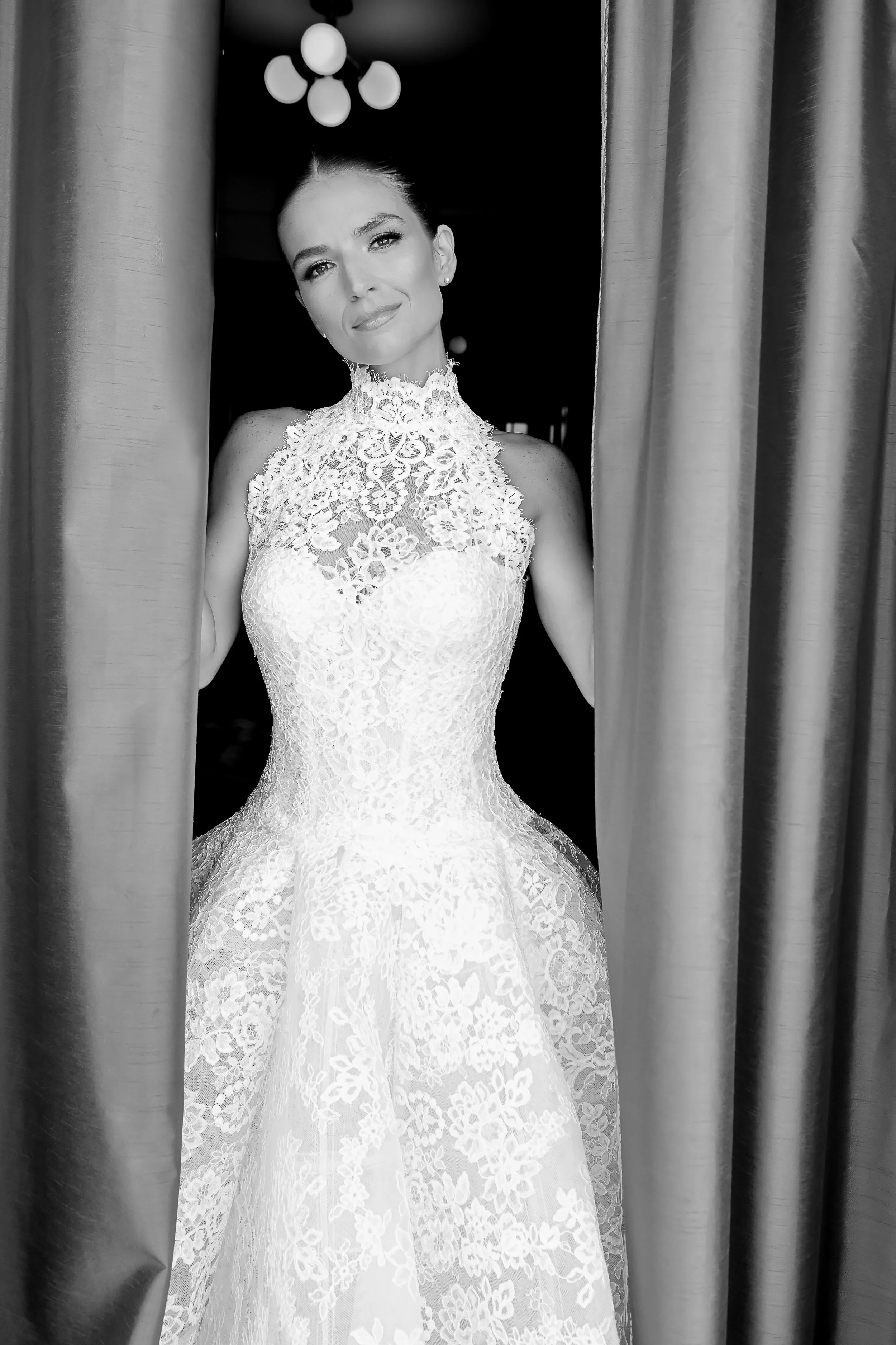A woman in an intricate lace wedding dress standing between curtains with a dark background overhead lights.