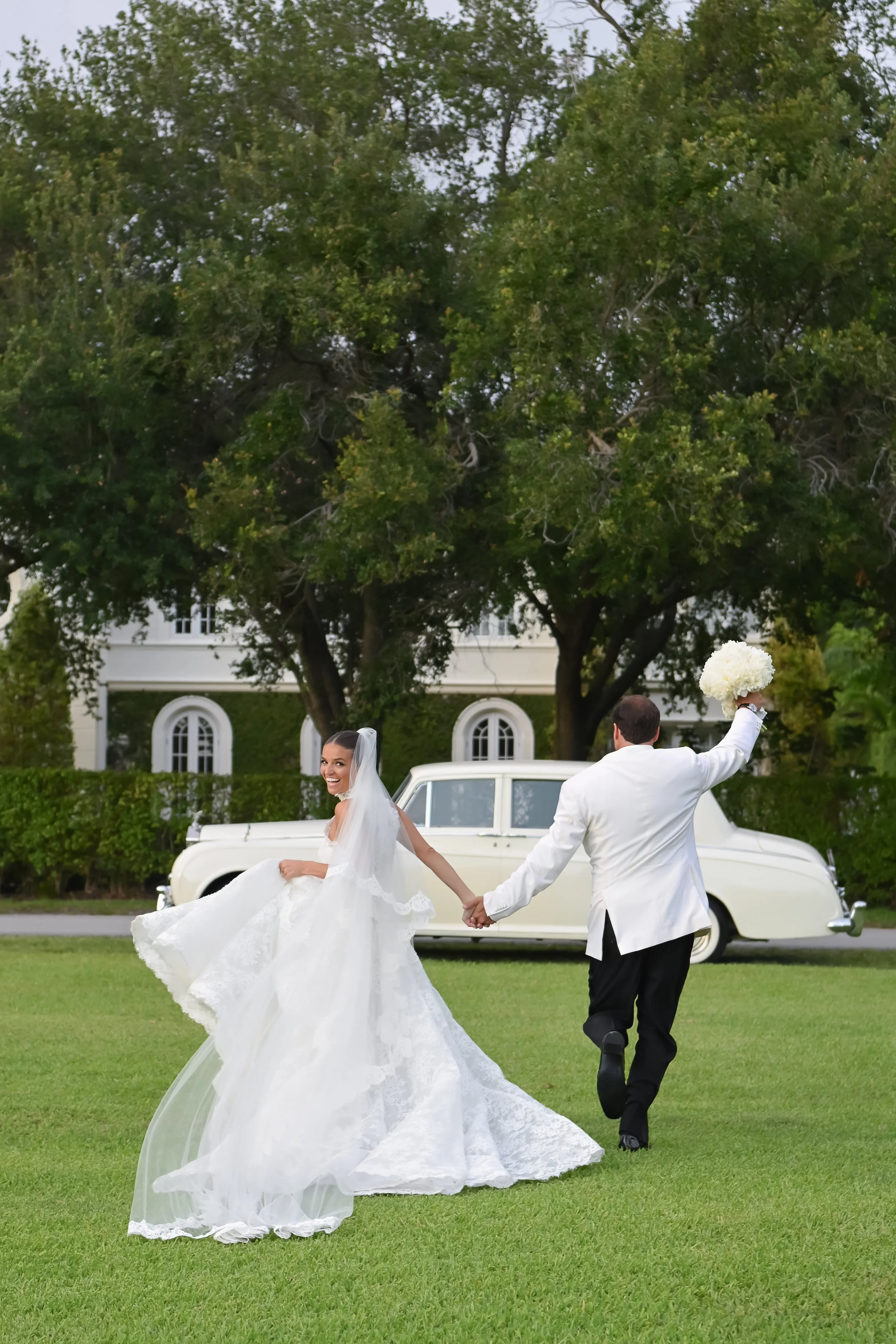 A bride and groom holding hands and running on a grassy lawn during their wedding celebration, with a vintage white car and large trees in the background.