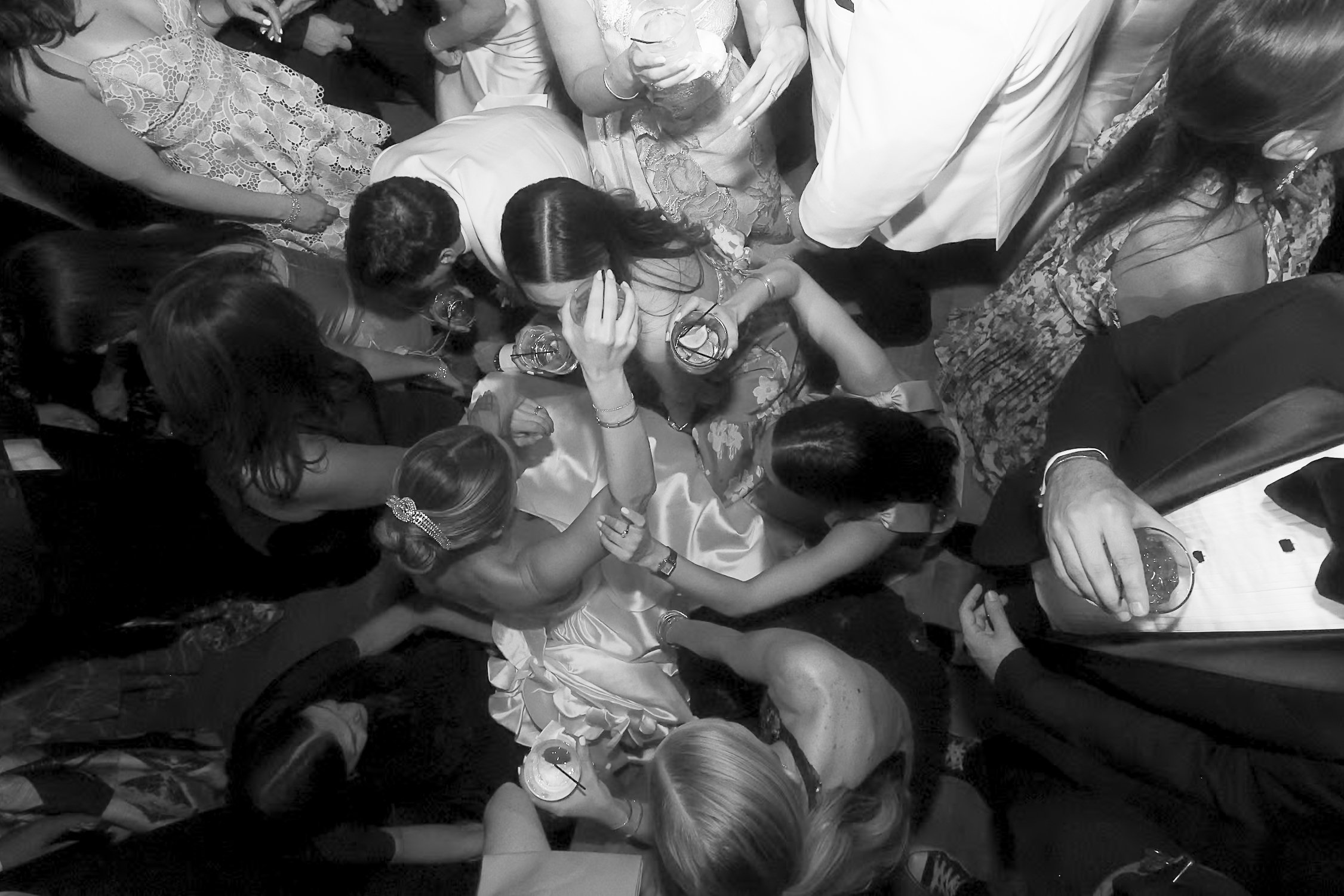 A black and white photo of numerous people at a party, with some holding drinks, some engaged in conversations, and others serving drinks at a bar or table. The focus appears to be on social interaction and celebration.