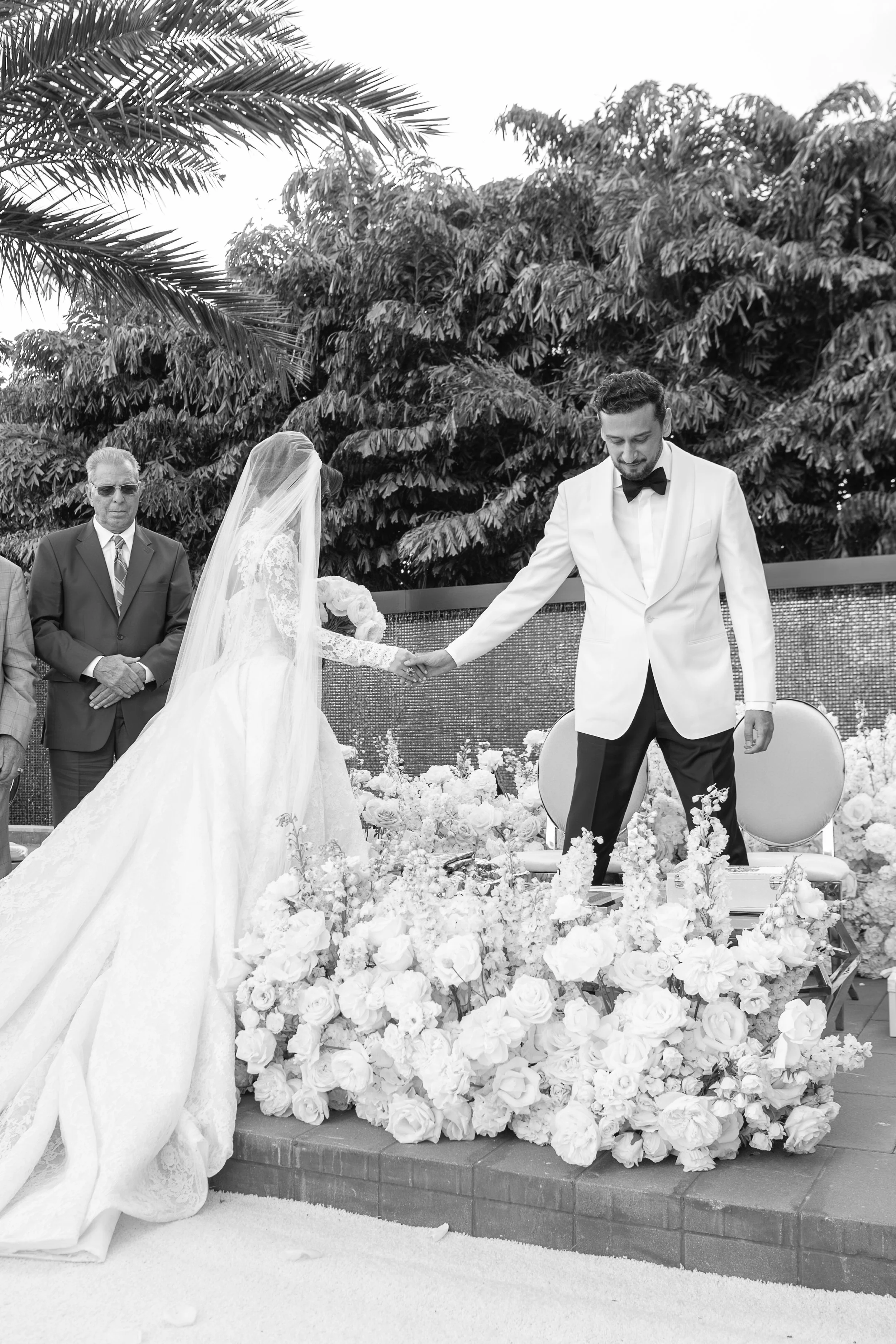 A black and white photo of a wedding ceremony with a bride and groom holding hands, surrounded by flowers, and officiants standing nearby. The scene is outdoors with lush foliage in the background.