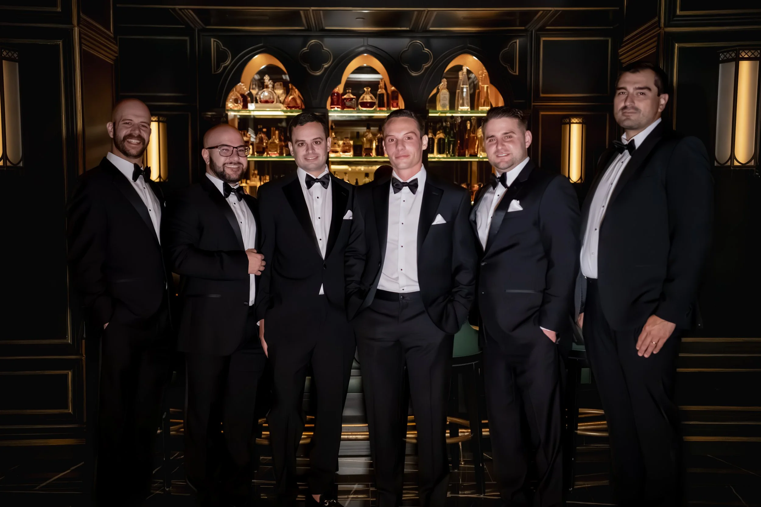 Group of six men in tuxedos standing together in front of a backlit bar with various bottles of alcohol in a luxurious setting.