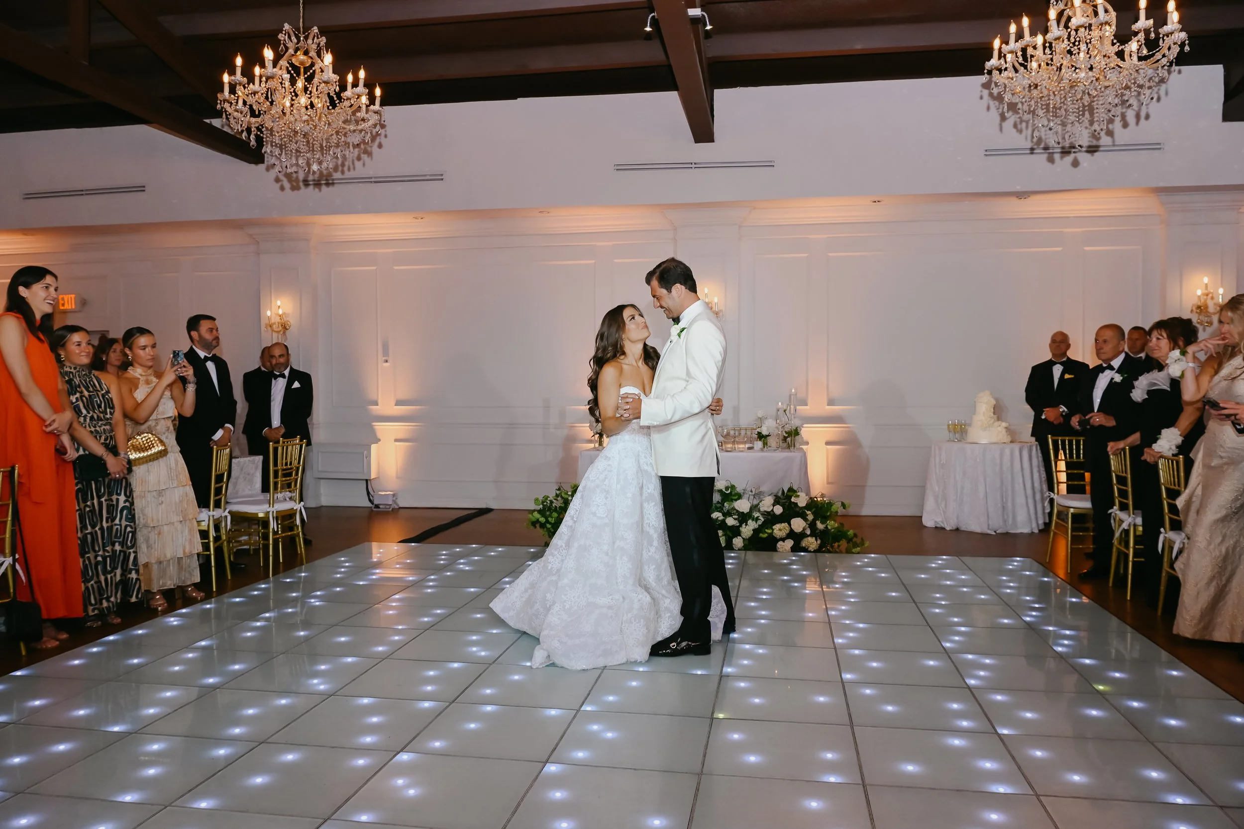 A bride and groom are dancing in the center of a wedding reception hall with guests gathered around watching. The hall is decorated with chandeliers, white walls, and floral arrangements. The dance floor has illuminated tiles, and there are tables wi