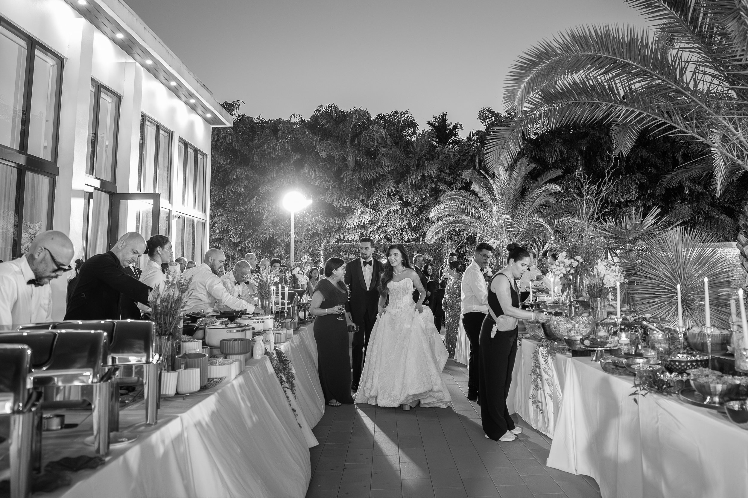 A wedding reception outdoors at night with a bride and groom walking past a buffet table. The reception area is decorated with flowers and surrounded by tropical trees and palm trees.
