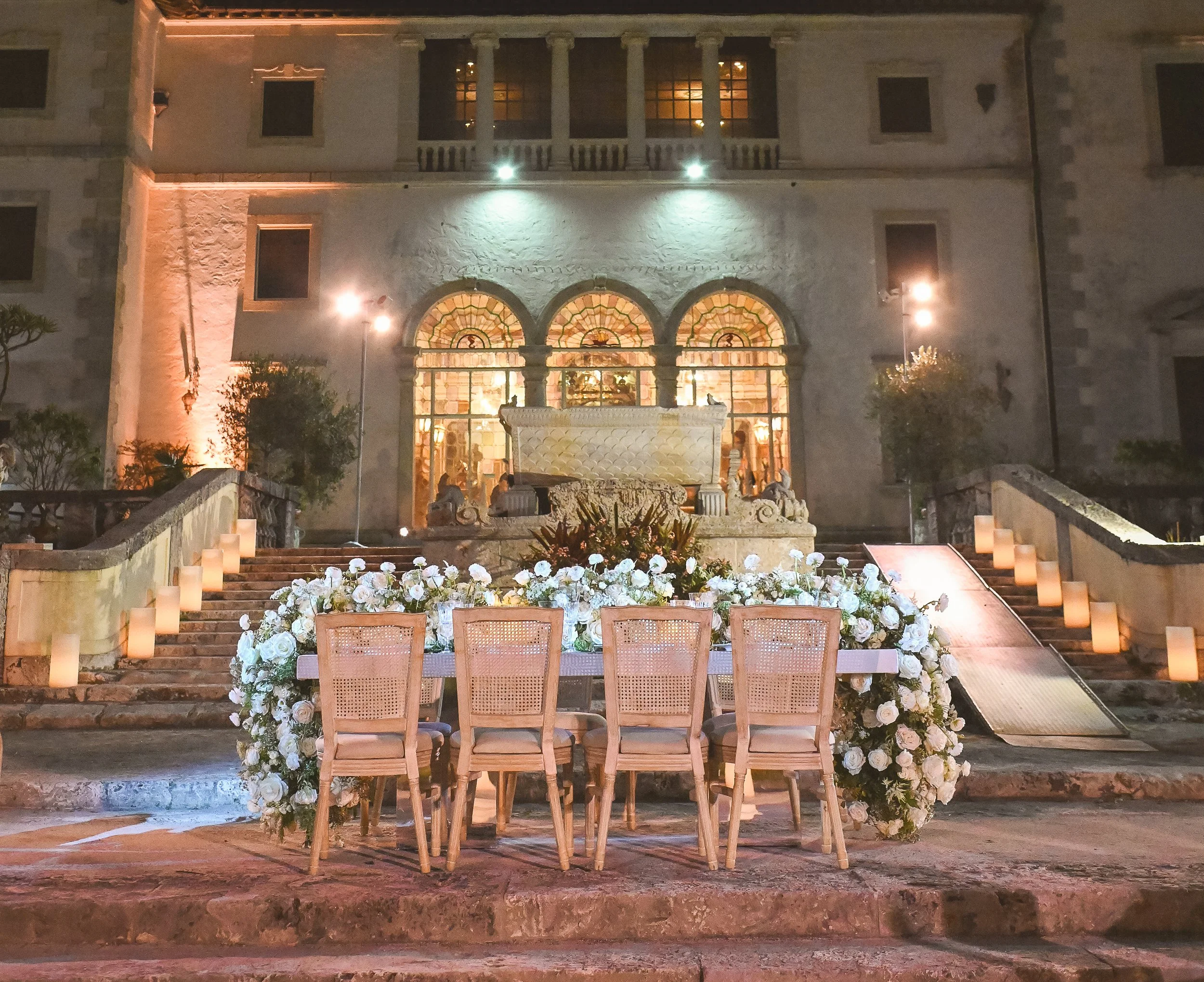 A staircase decorated with a large arrangement of white roses and greenery leads to an illuminated building with a stone facade, arched windows, and a fountain at the bottom of the stairs, set for an evening event.