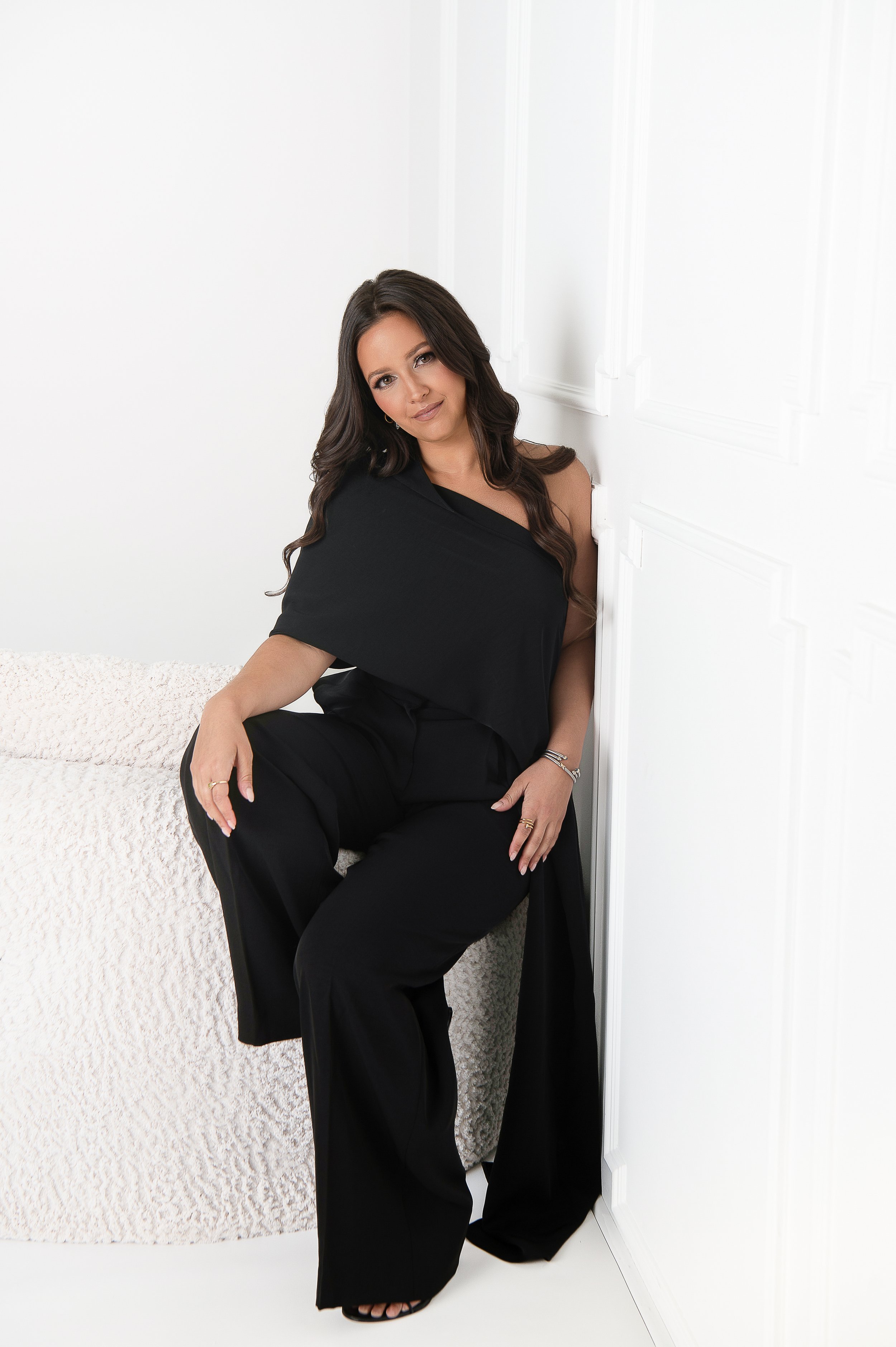 A woman with long dark hair wearing a black dress and black pants, sitting on a beige textured surface, leaning against a white wall and corner, looking at the camera with a slight smile.